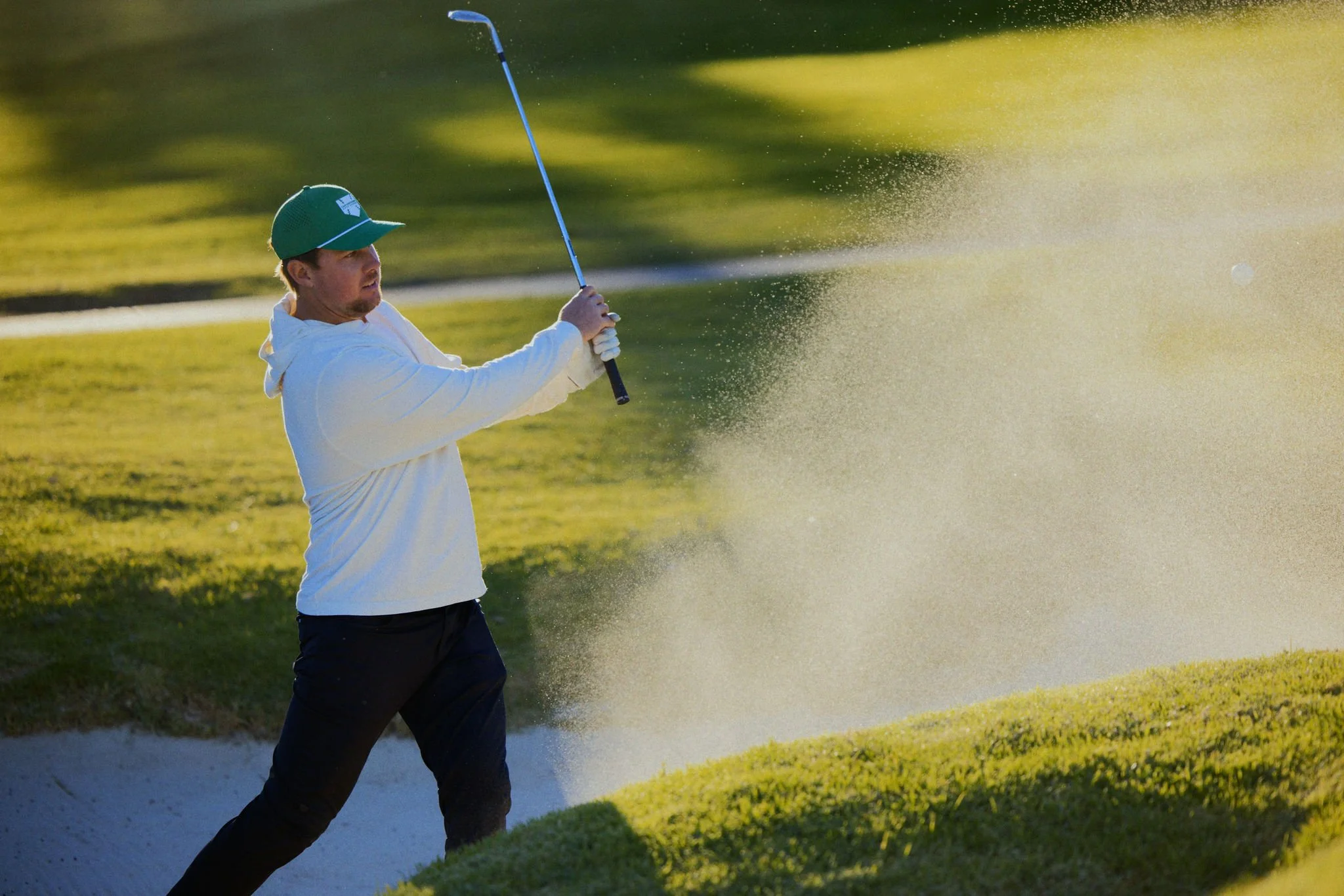 A man in a white hoodie and black pants playing golf, hitting a shot from a sand trap on a golf course in the late afternoon or early evening sunlight.