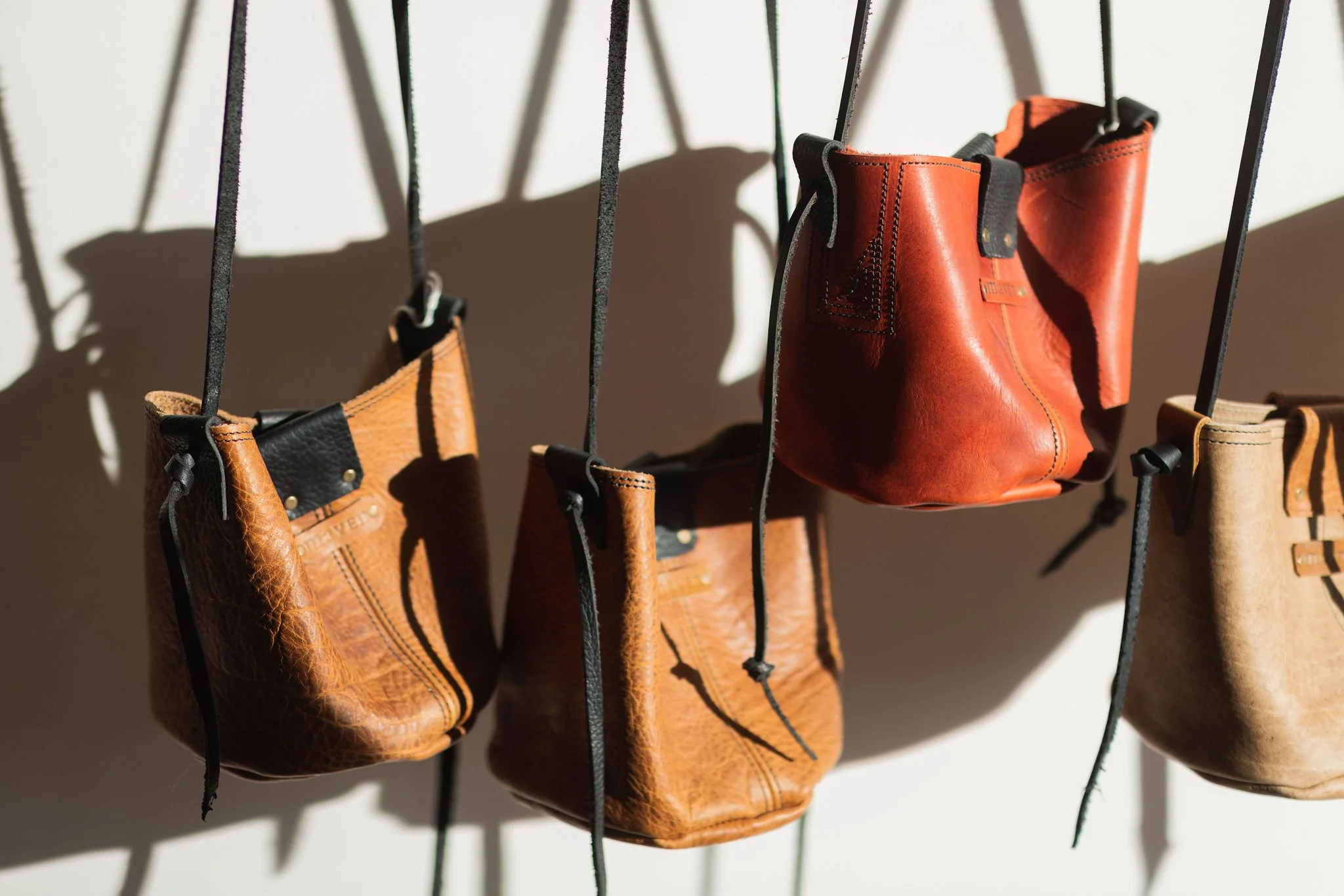 Assorted leather bags hanging from straps with shadows on a wall.