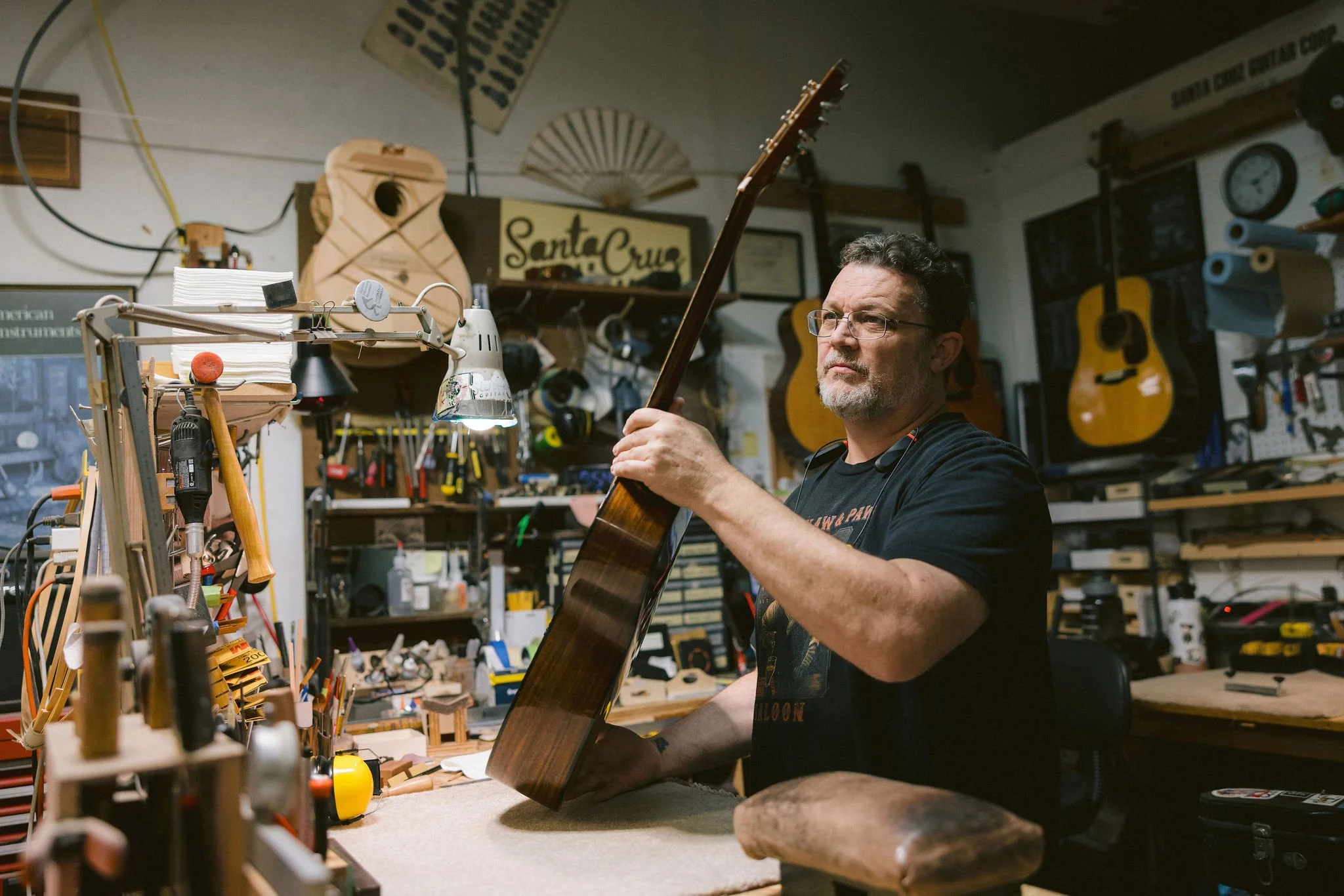 A man in a workshop is holding a guitar neck and examining it. The workshop is filled with tools, guitars, and musical equipment.