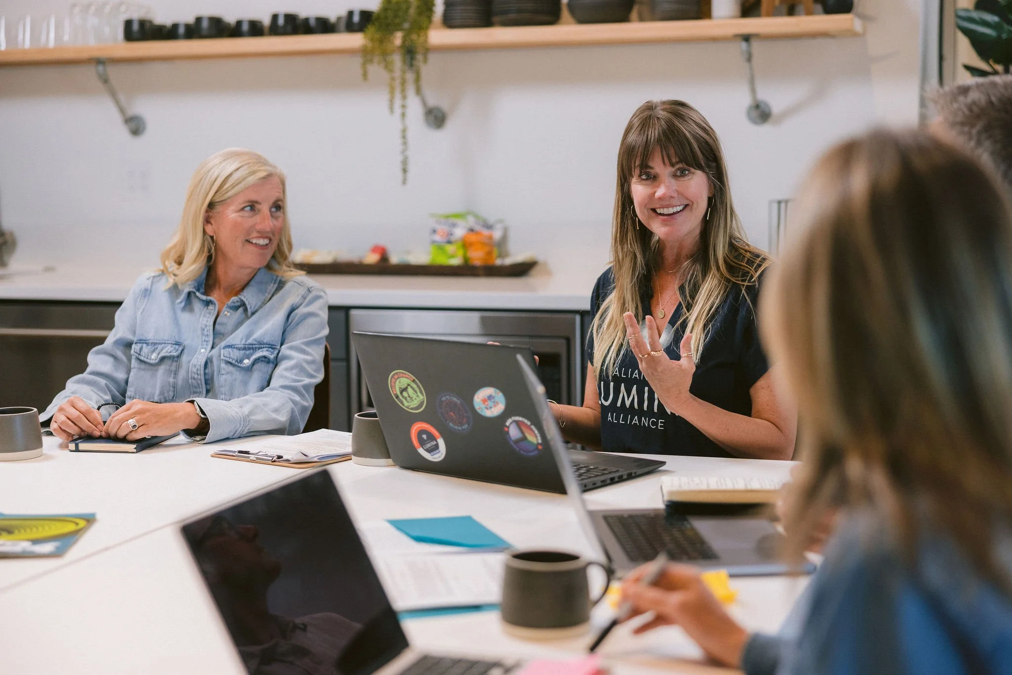 A group of women sitting around a white table in a meeting or discussion, with one woman actively speaking and smiling, using a laptop, in a modern kitchen or office space.