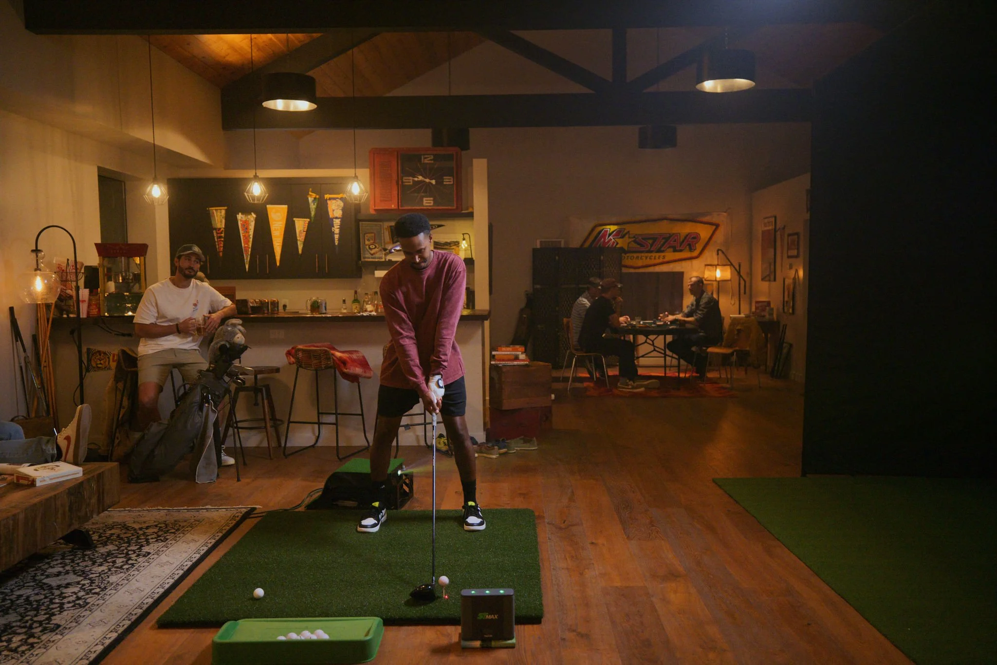 Indoor scene with people in a cozy room. One person is playing golf on a small indoor putting green golf simulator, while others are sitting at a table in the background. The room has warm lighting, wooden floors, and various decorations.