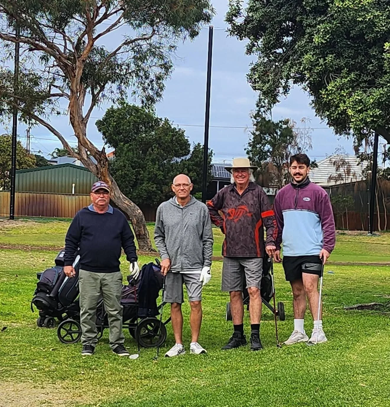 Four men standing together on a golf course, some holding golf clubs and a golf push cart, with trees and a fence in the background.