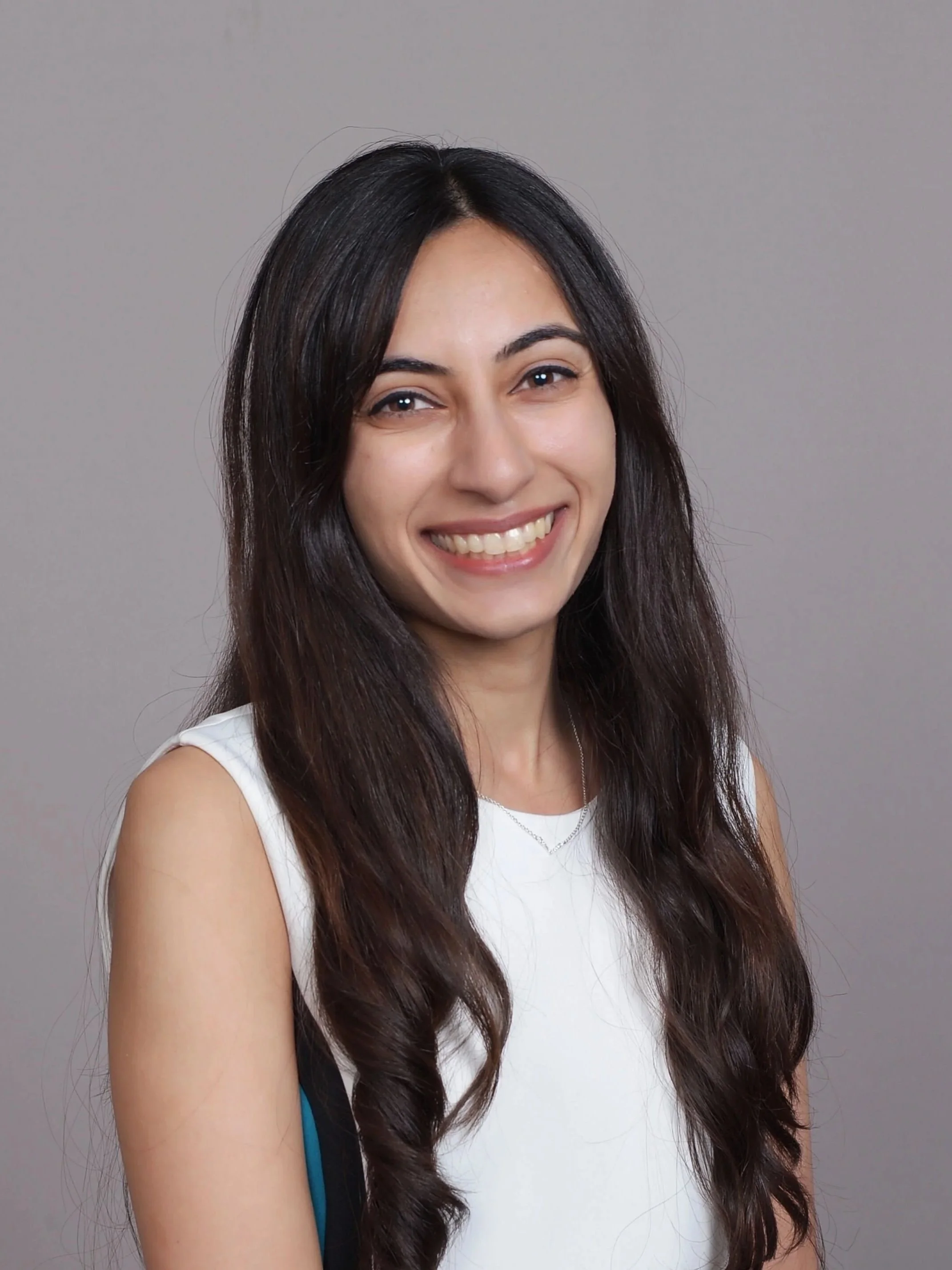 Smiling woman with long wavy dark hair wearing a blue and white checkered shirt and a delicate necklace, standing against a plain gray background.