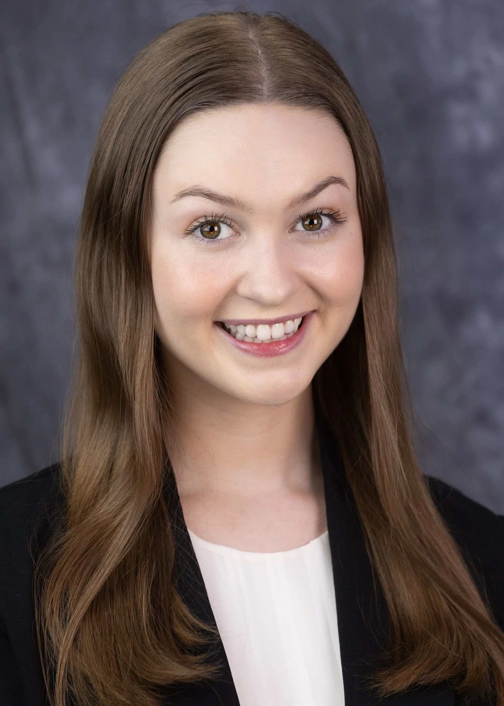 A young woman with long light brown hair, wearing glasses, a white shirt, and a black blazer, smiling against a plain grey background.