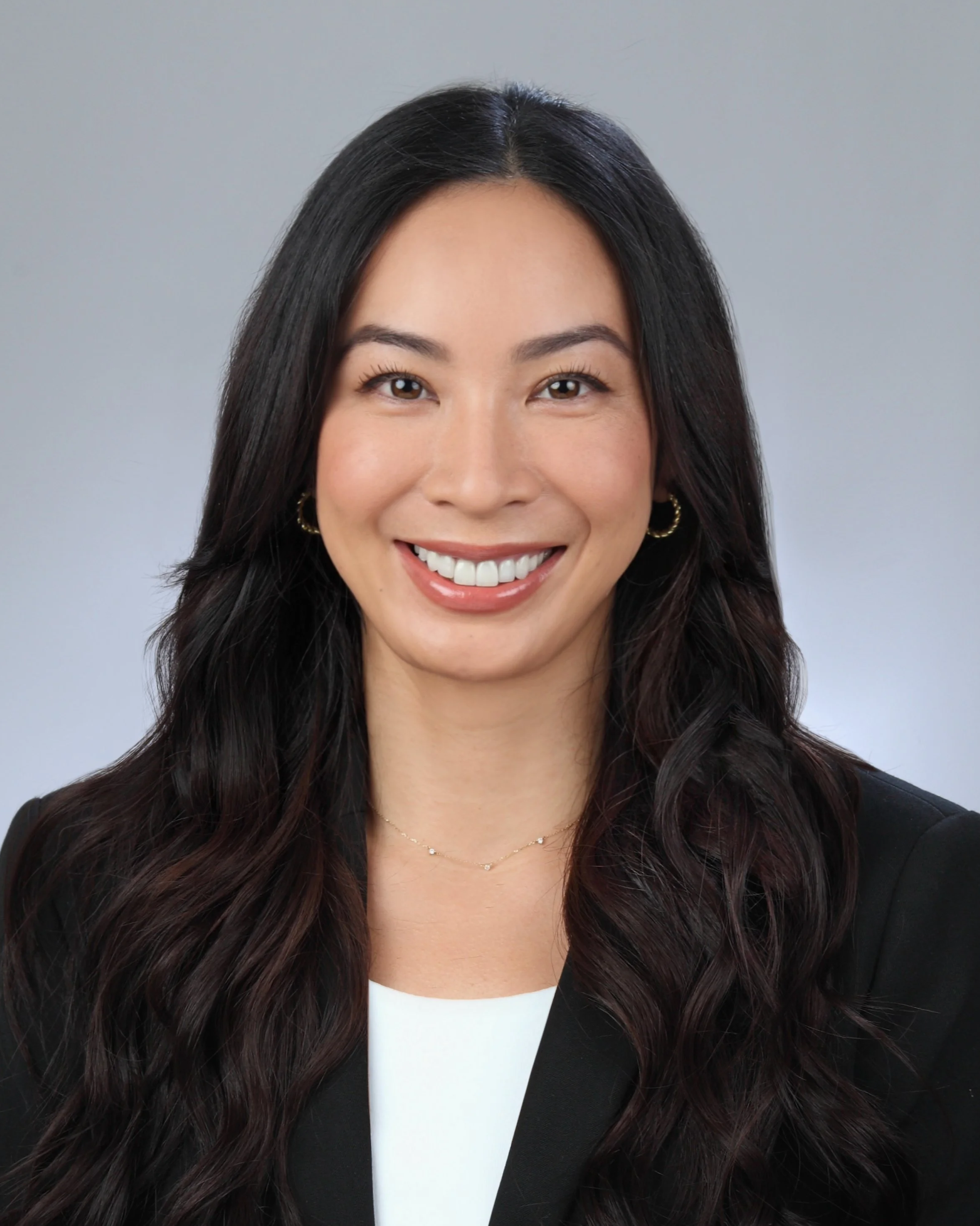 Portrait of a smiling woman with long brown hair, wearing a black blazer and white top, against a gray background.