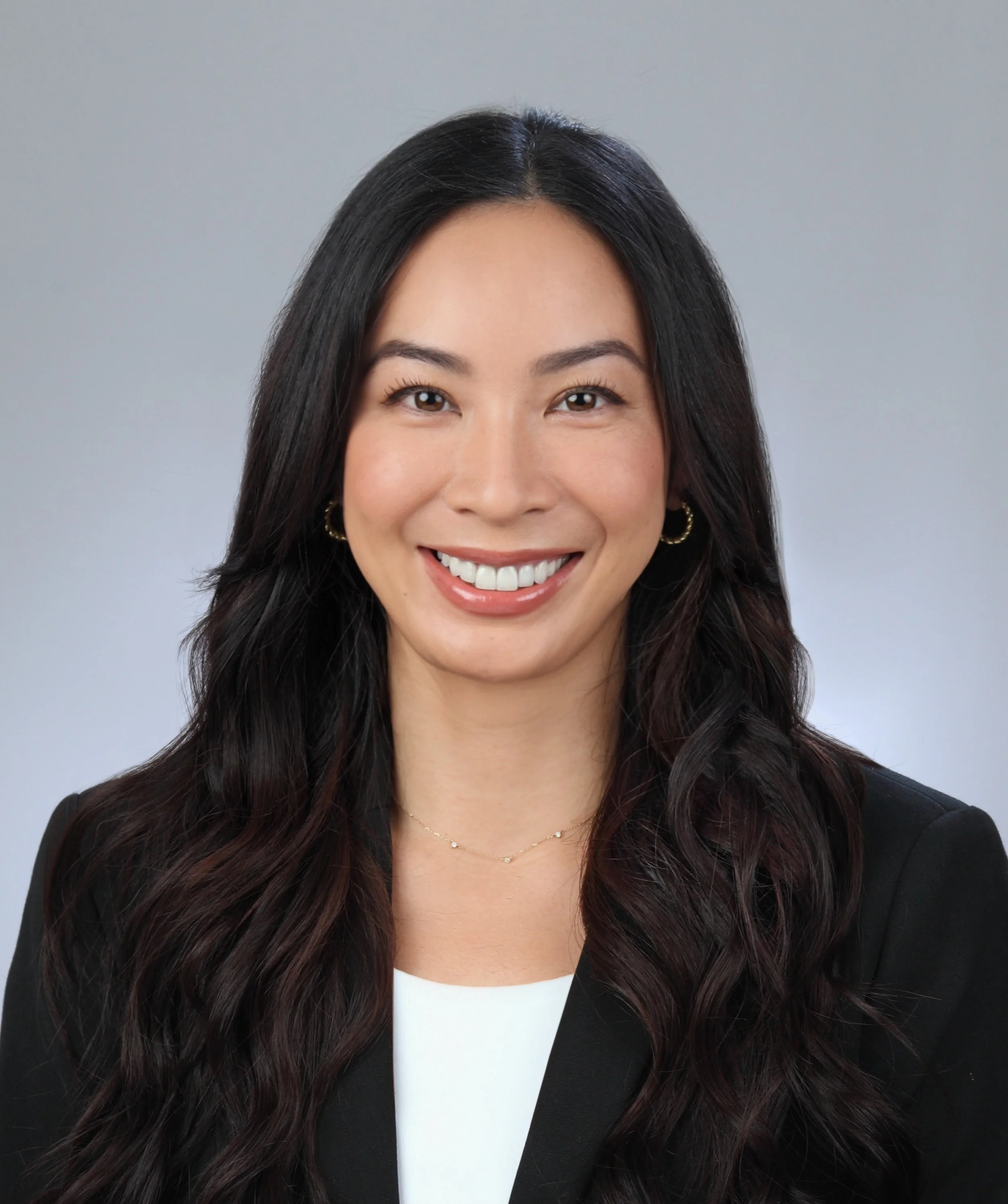Portrait of a smiling woman with long brown hair, wearing a black blazer and white top, against a gray background.