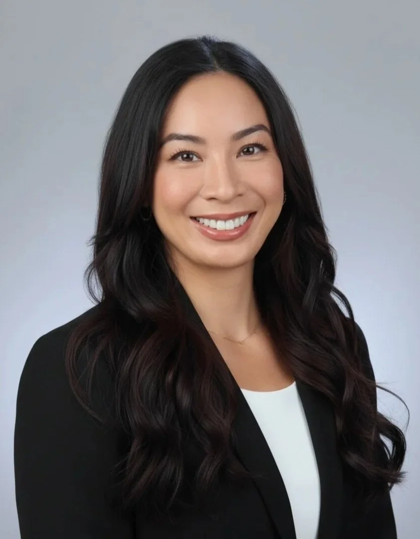 Portrait of a smiling woman with long brown hair, wearing a black blazer and white top, against a gray background.