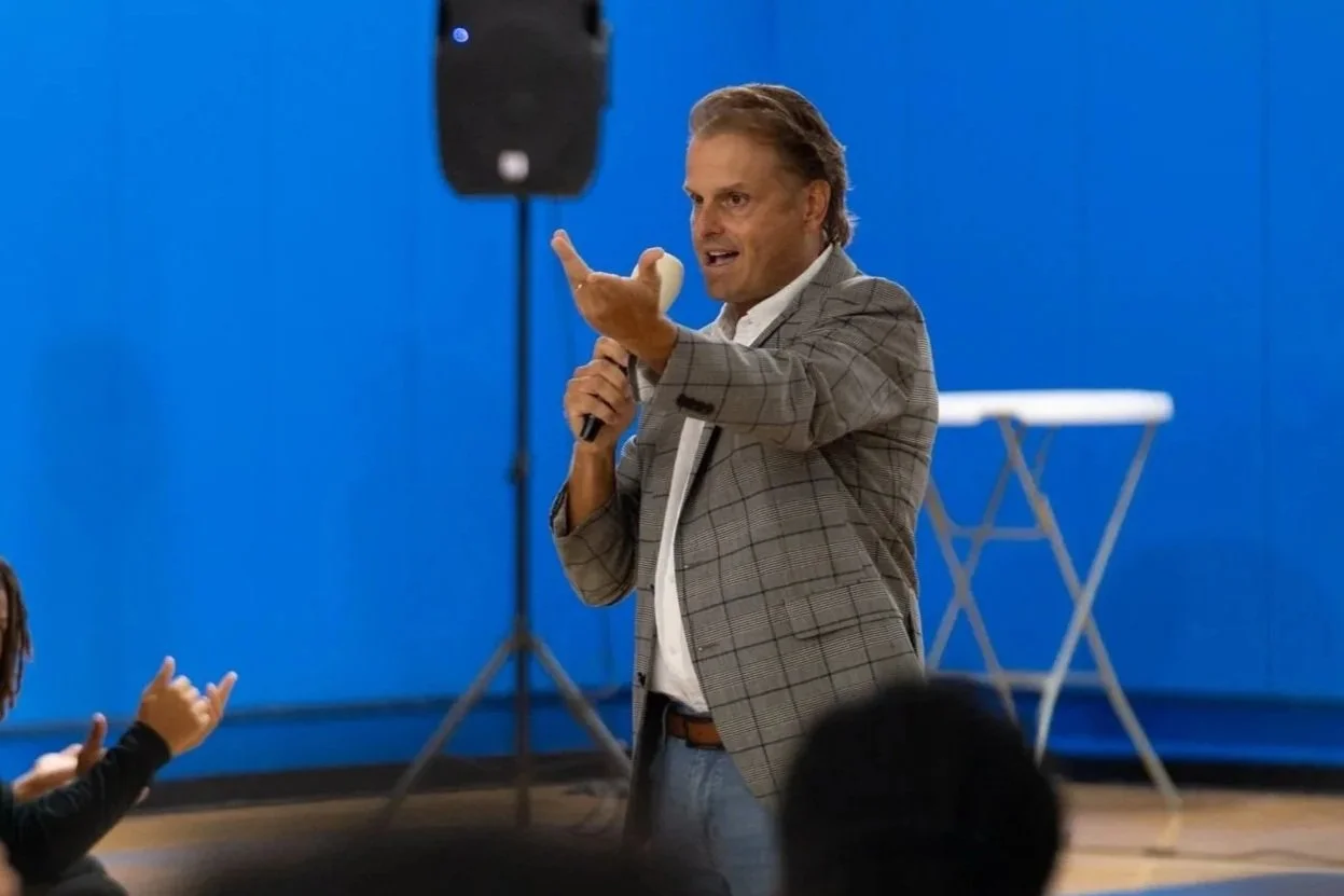 A man in a checkered blazer speaking into a microphone during a presentation or speech, with a blue wall in the background and a white folded table behind him.