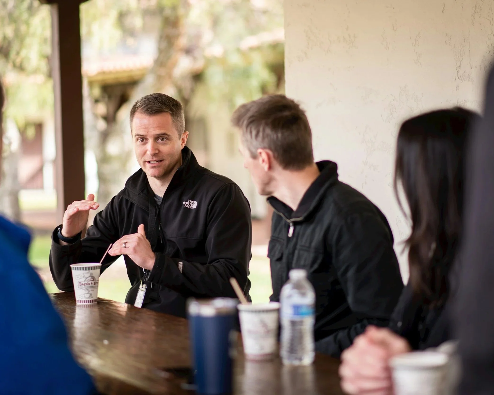 A man gesturing and speaking during a conversation with three people seated at a wooden outdoor table, with cups and bottled water in front of them, and blurred trees and building in the background.