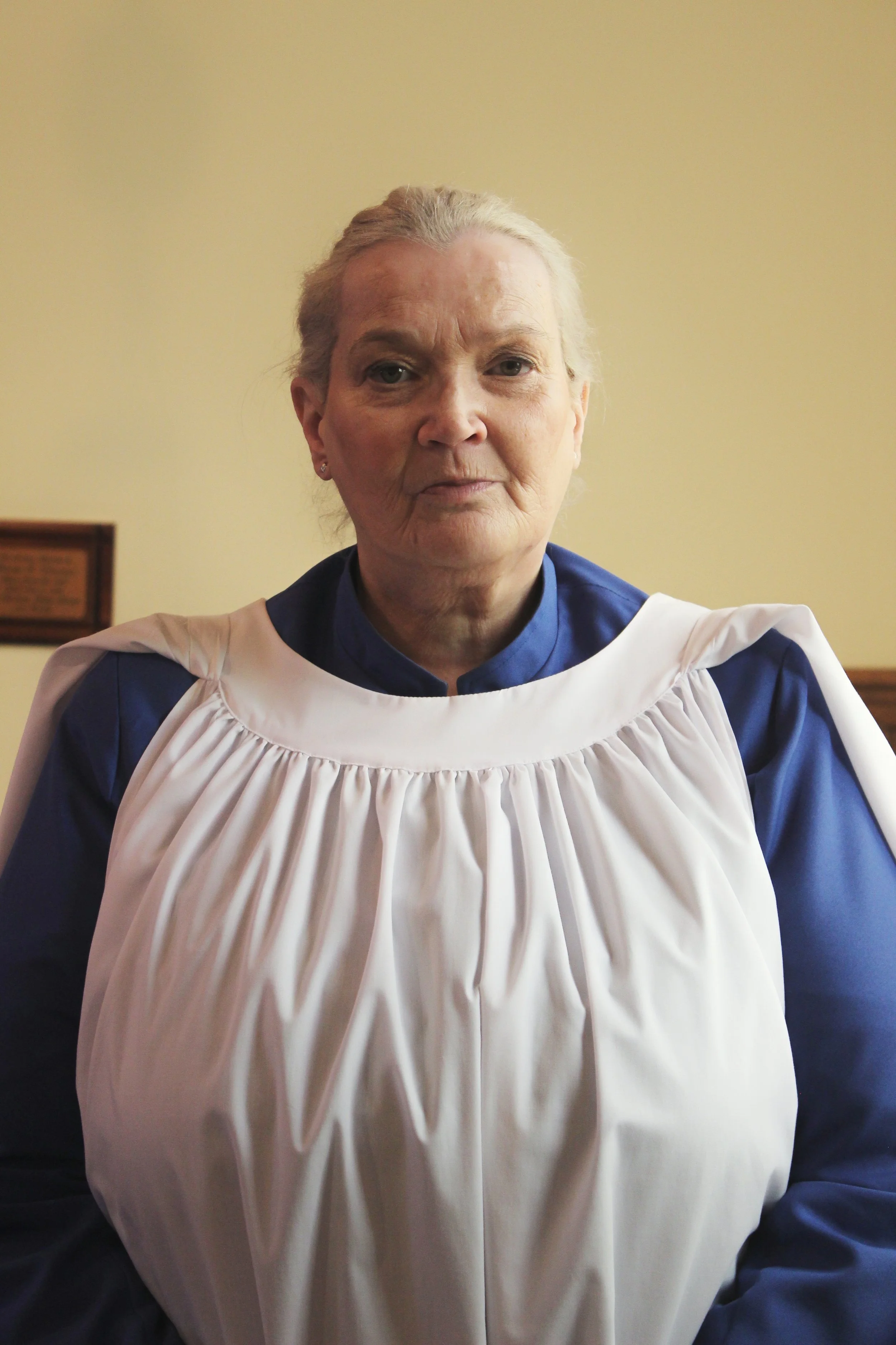 An older woman wearing a blue uniform with a white religious or ceremonial collar or apron, standing in a room with a plain light-colored wall in the background.