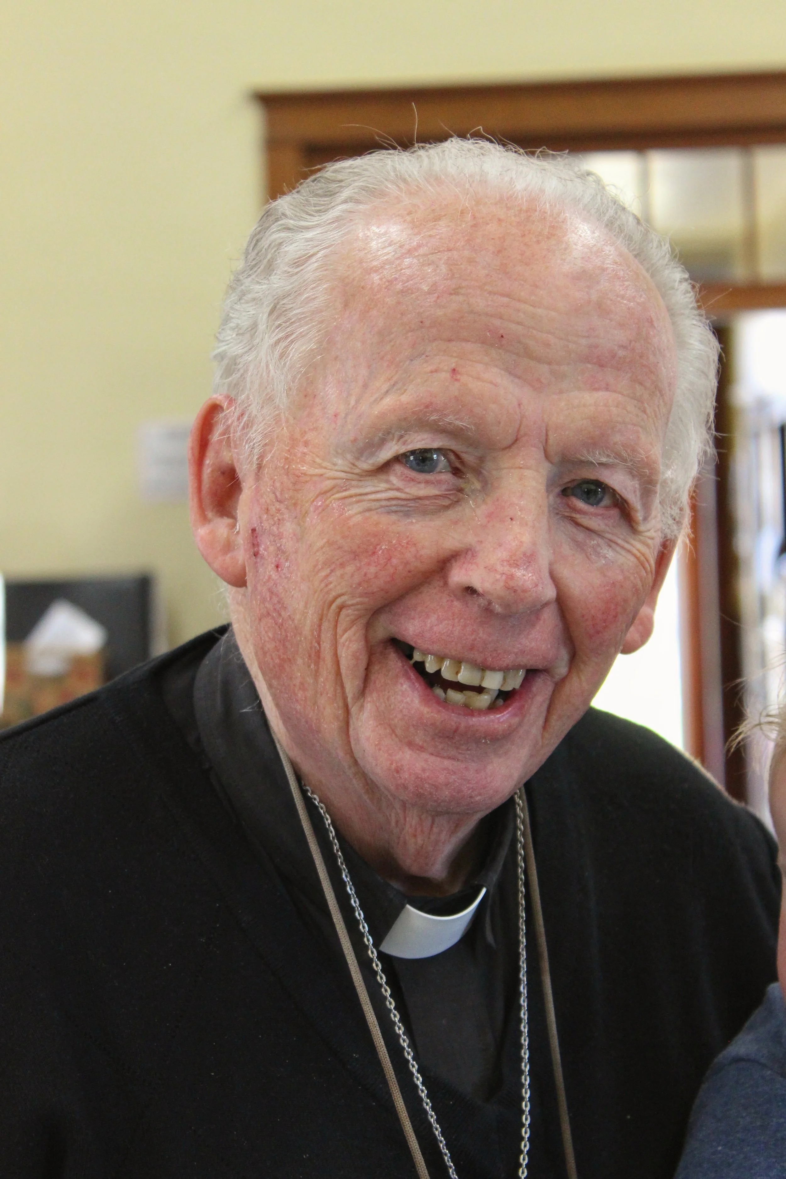 Portrait of an elderly man with a broad smile, wearing a clerical collar and a black shirt, indoors near a wooden doorframe.