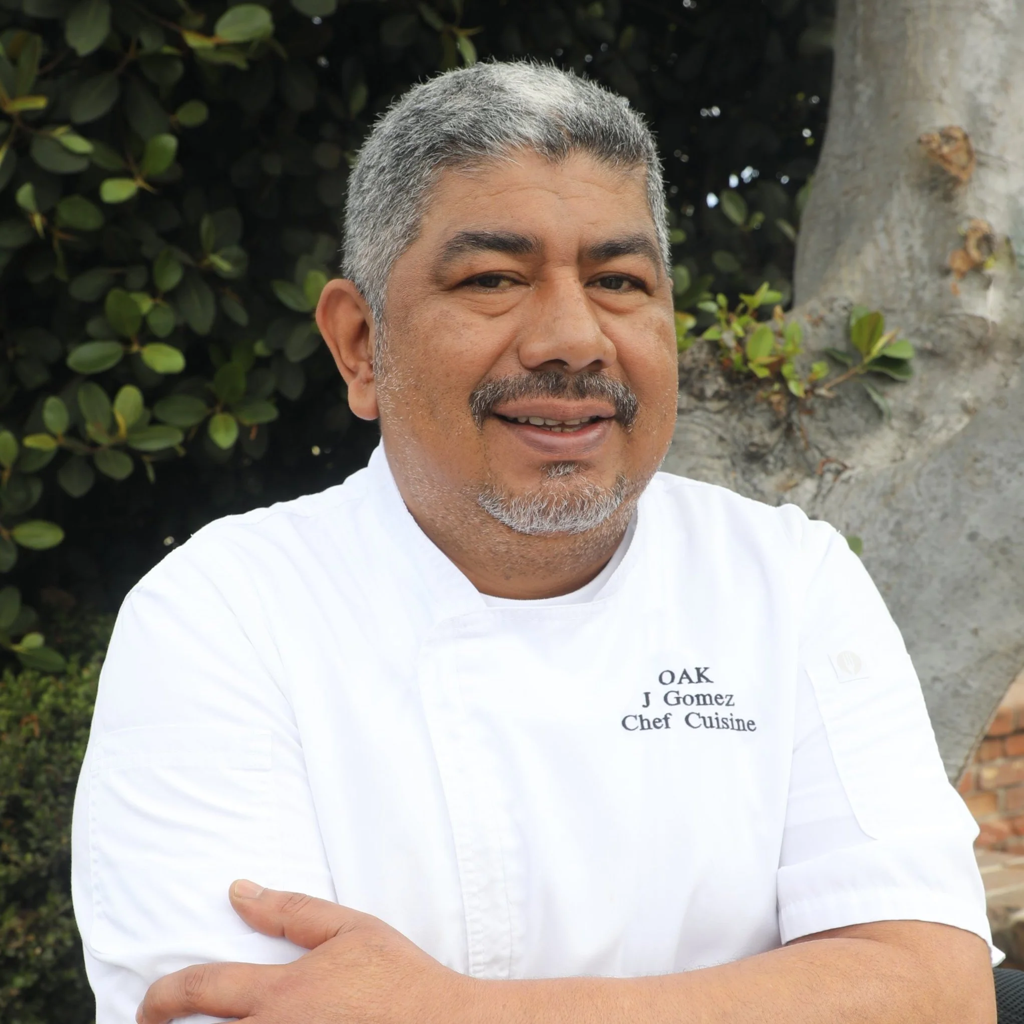 Our Chef, Jorge, who is hispanic, wearing a white chef's coat. He is sitting in front of a green hedge and tree.