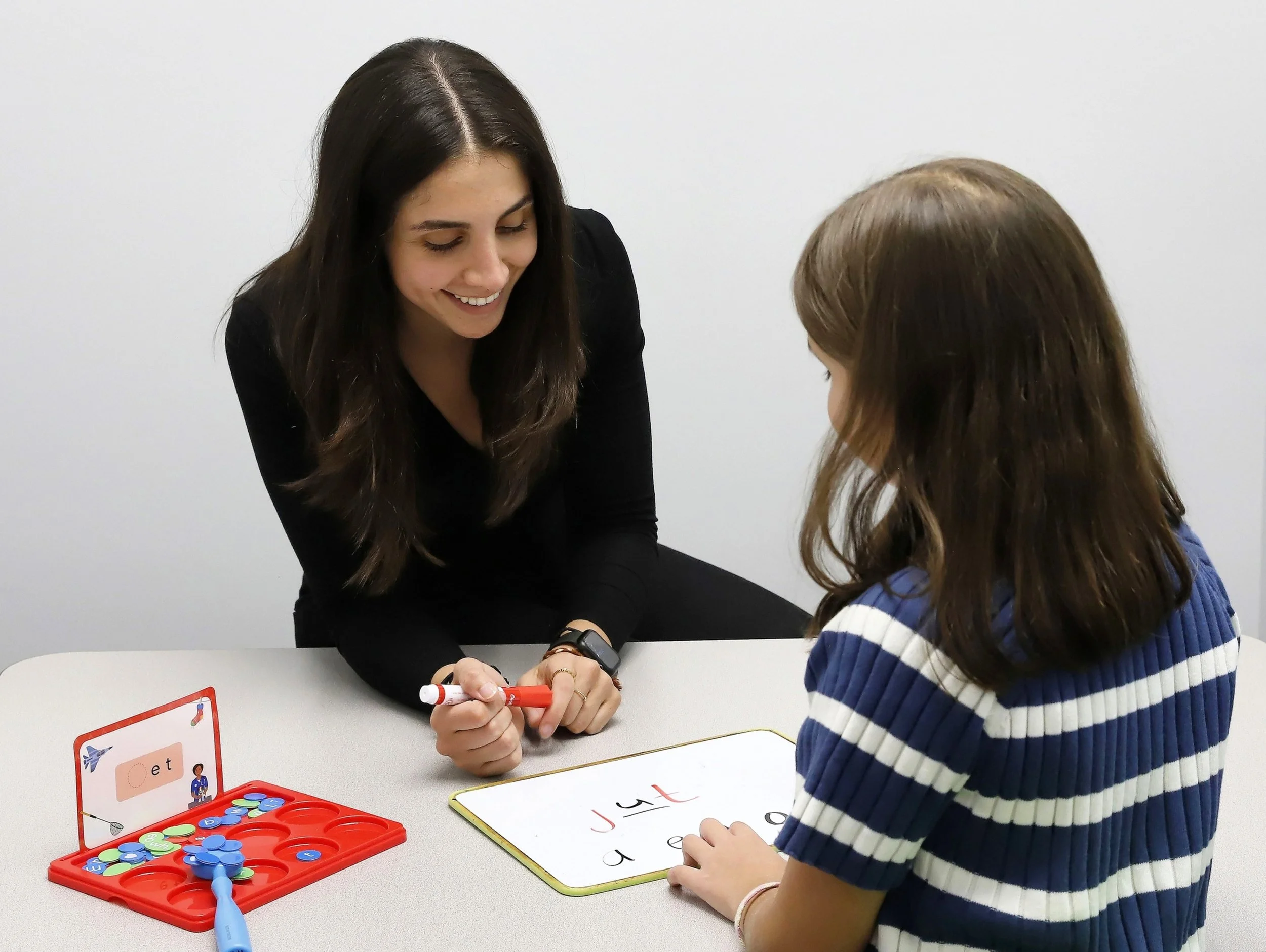 A speech-language pathologist working one-on-one with a young girl on spelling and literacy skills using a whiteboard and letter tiles