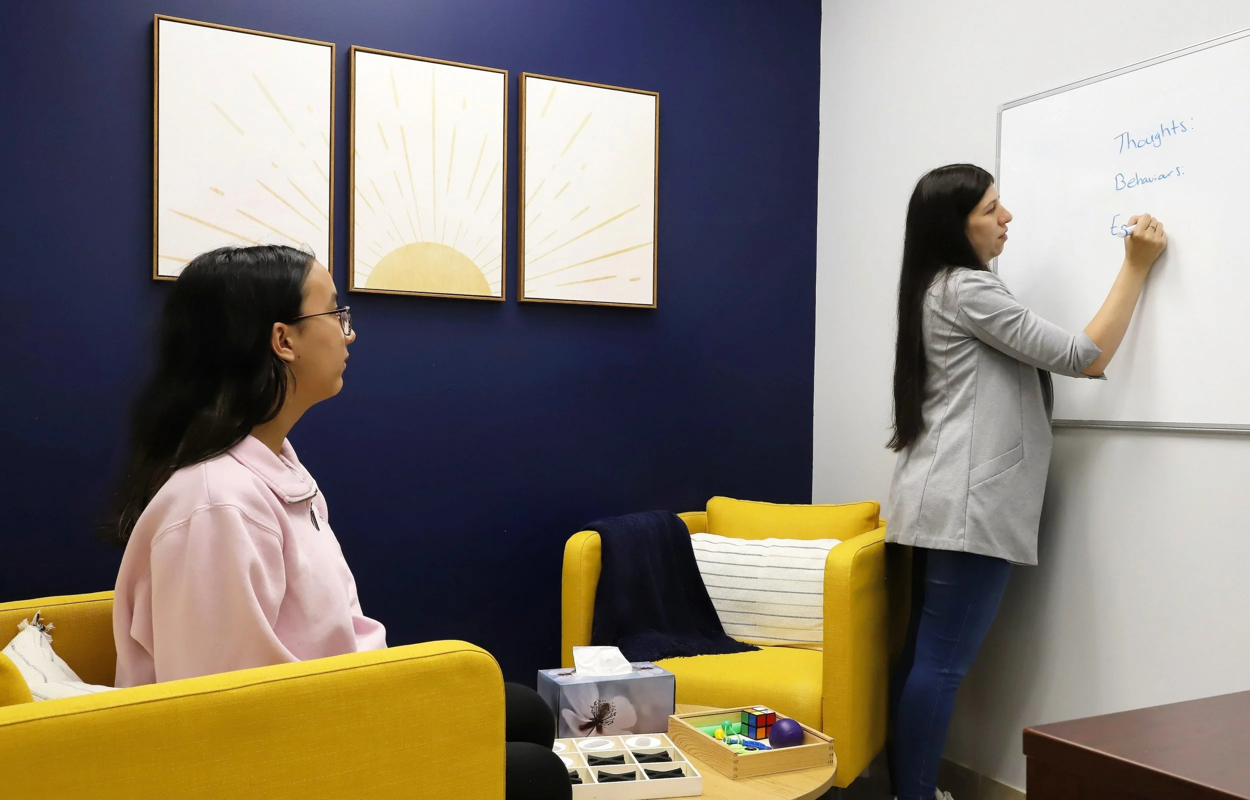 A therapist writing thoughts, behaviours, and emotions on a whiteboard during a CBT session with a teenage client in a warm, welcoming therapy room