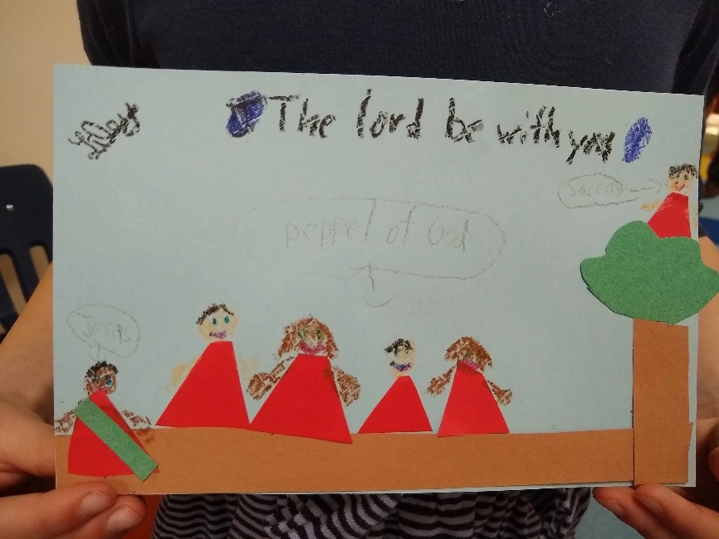 A handmade card featuring simple drawings of five children with different hair colors, all wearing red outfits, and a large green tree with a brown trunk. There are handwritten messages saying 'The Lord be with you', 'Shalom', and 'Peace'. The card is held by children with striped shirts.