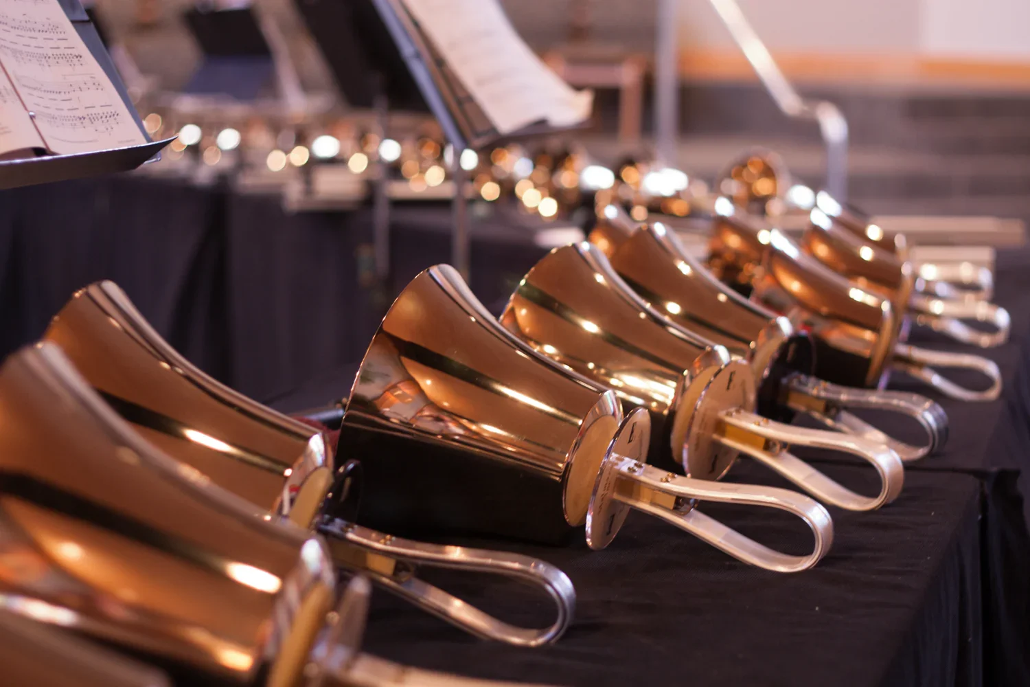 A row of shiny brass bells on a black table, with sheet music and music stands in the background.