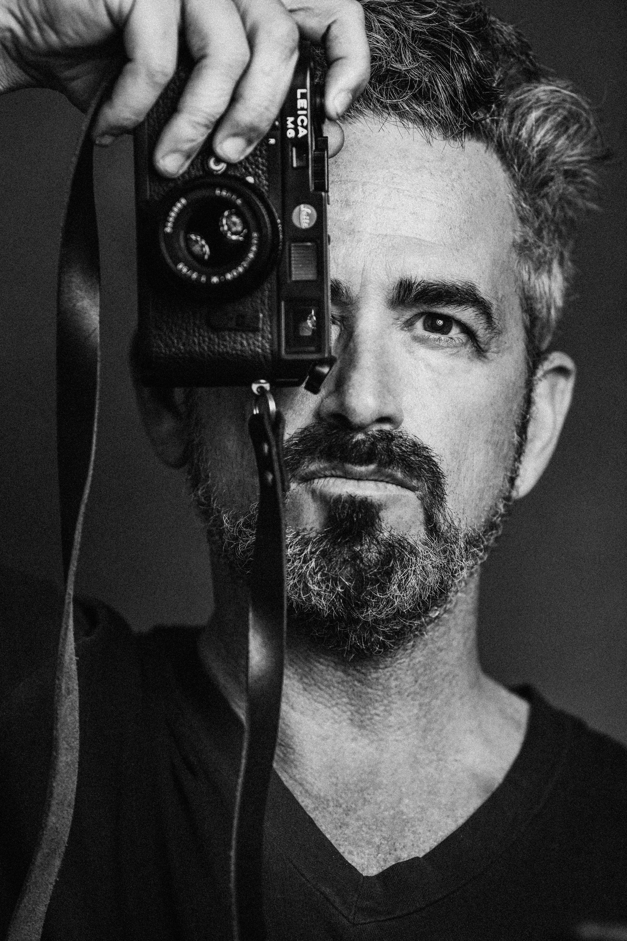 A man with a beard holds up a camera covering on eye in a self portrait style photograph.