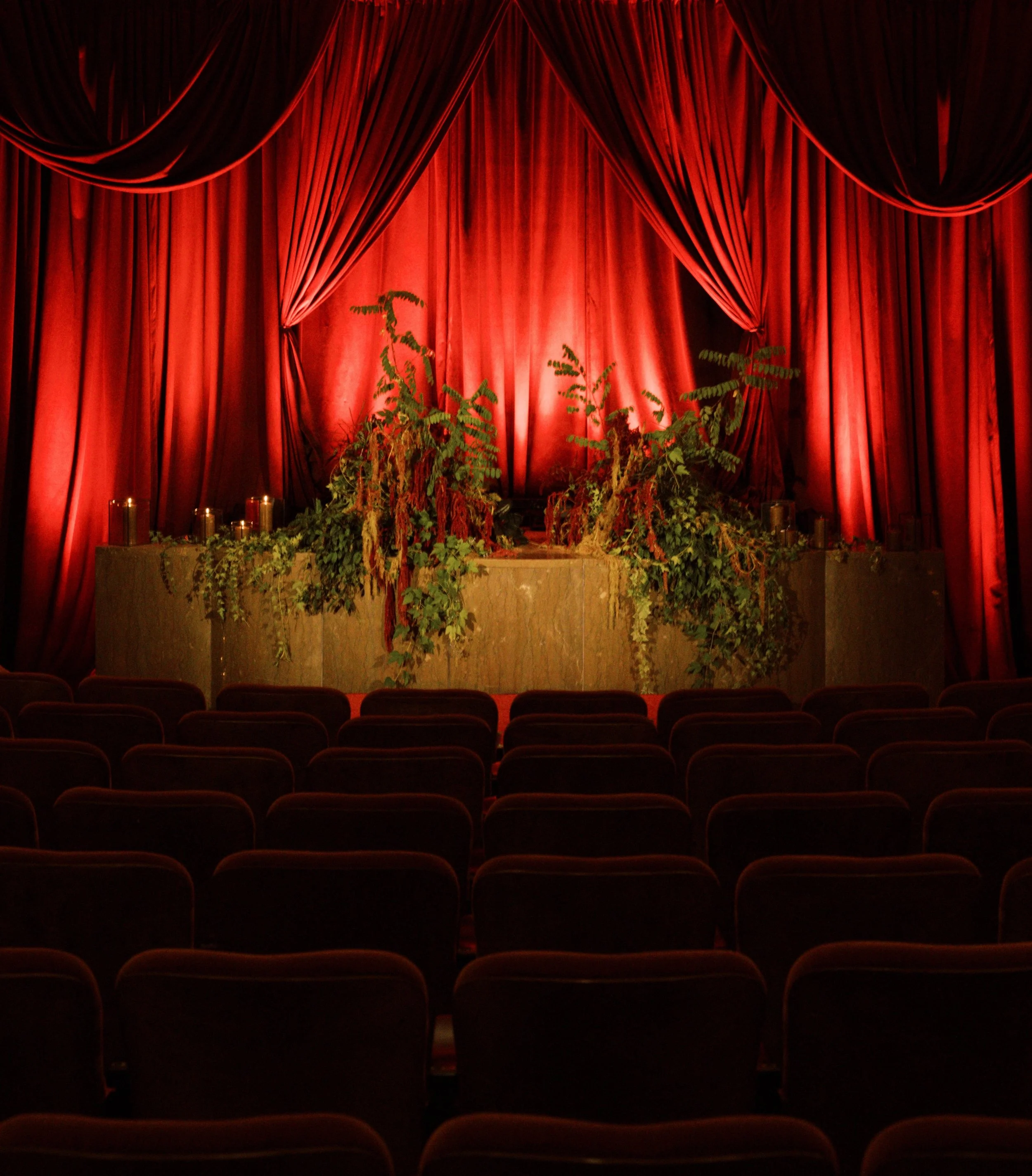 Theater stage set for a luxury California wedding reception with deep red curtains, greenery, candles, and guest seating.