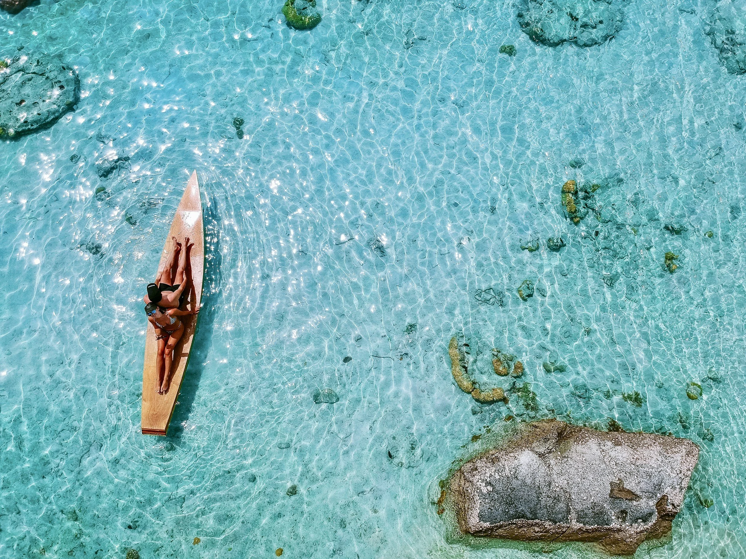 A person lying on a surfboard in clear, blue water with rocks and coral beneath the surface.