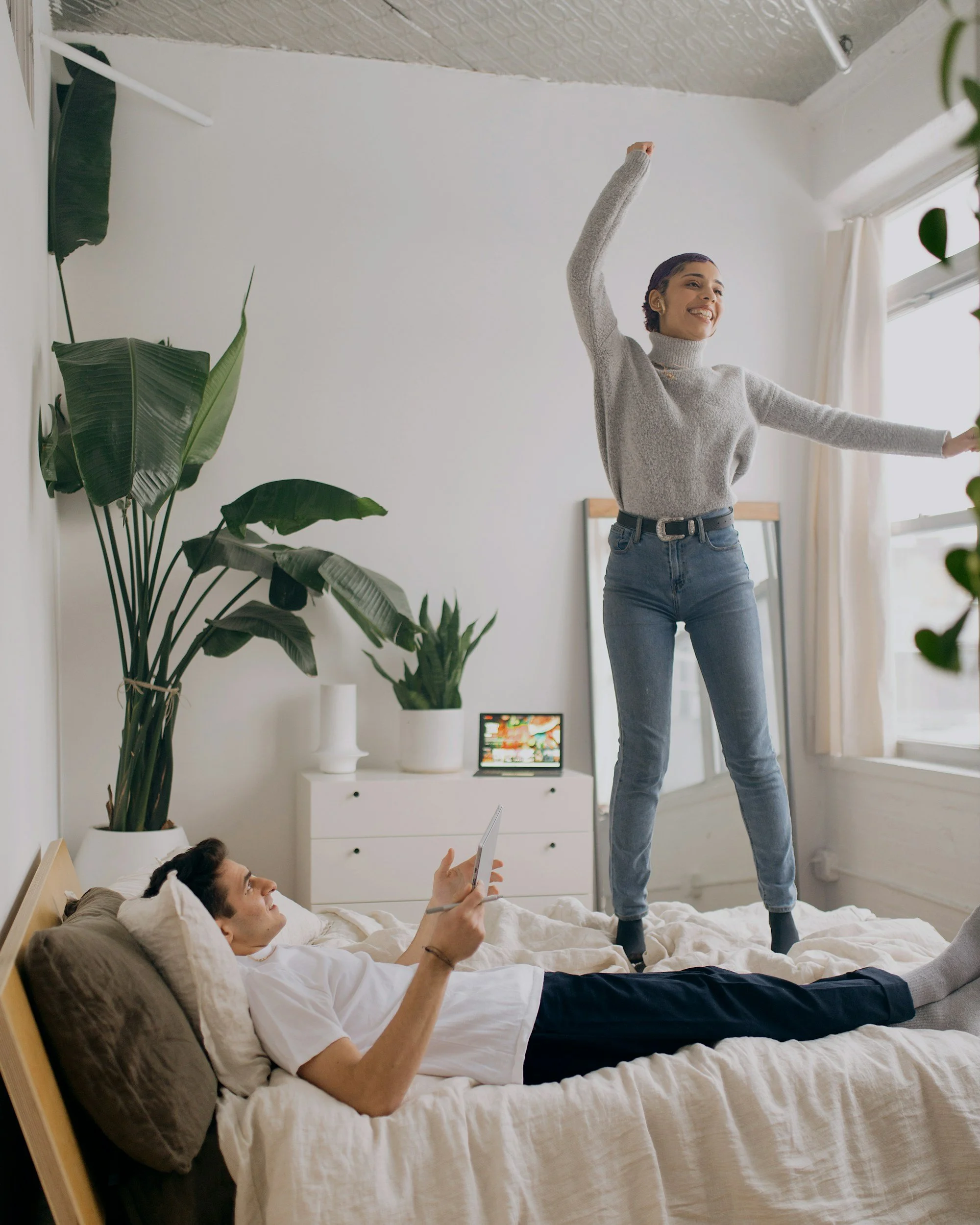 A woman and a man are on a bed in a bright, modern bedroom. The man is lying down, holding a tablet, wearing a white t-shirt and dark pants. The woman is standing on the bed, wearing a grey turtleneck sweater and jeans, smiling with arms raised. There are large potted plants, a small TV, and a window with curtains in the background.