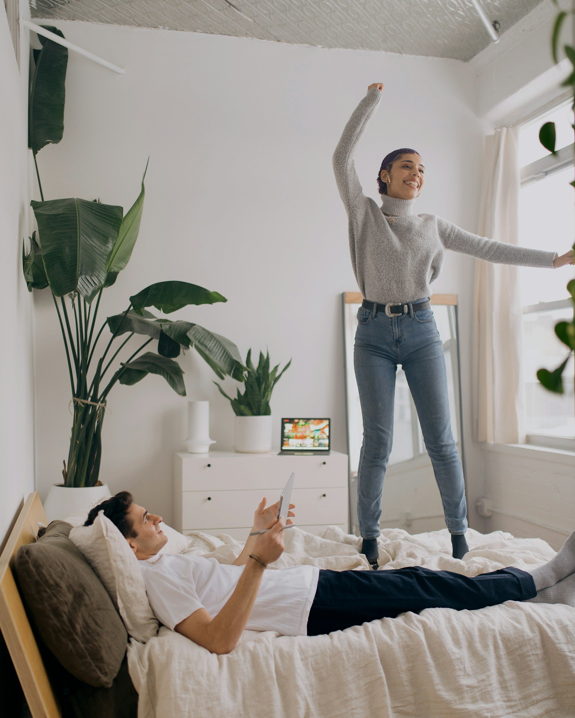 A woman with dark, curly hair is smiling and standing on a bed, stretching her arms out. A man is lying on the bed below her, looking up and holding a tablet.