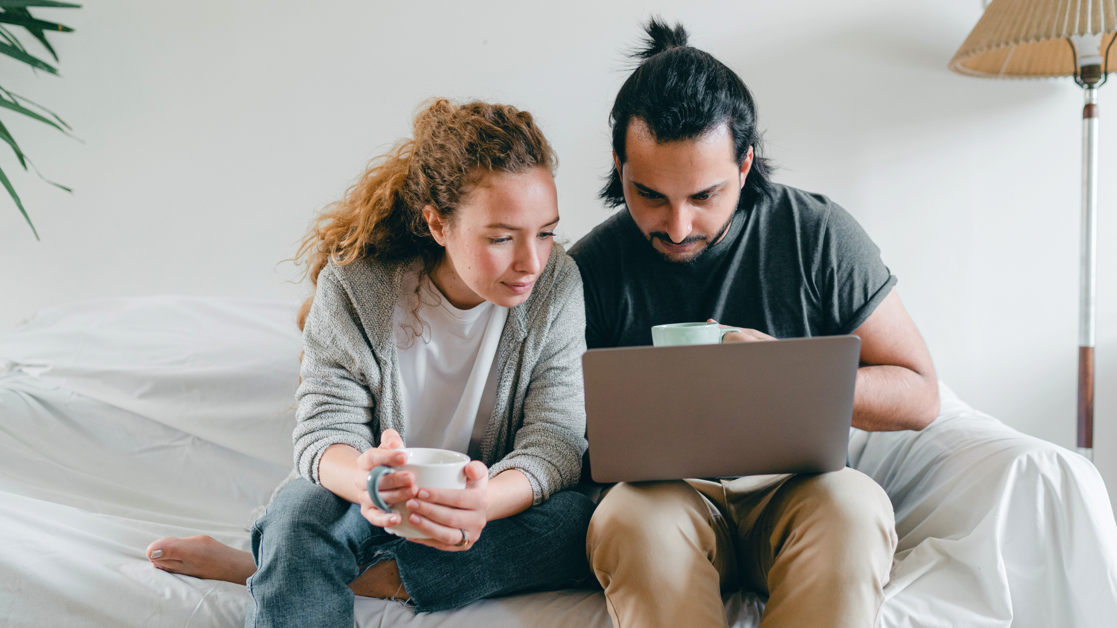 A man and woman sitting on a bed, looking at a laptop together, holding coffee mugs