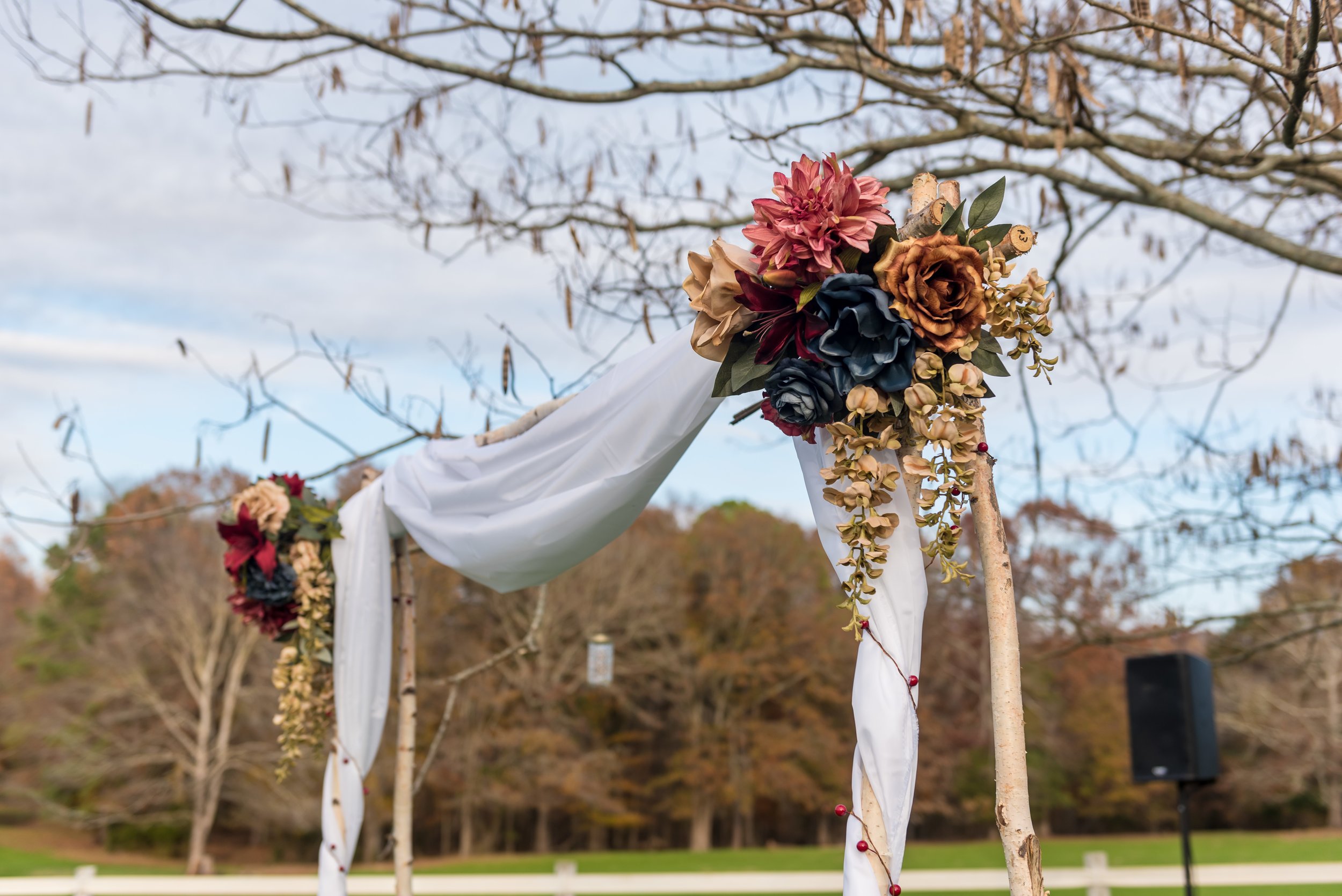 Wedding arch decorated with flowers and white fabric, set in an outdoor park with trees and a speaker in the background.