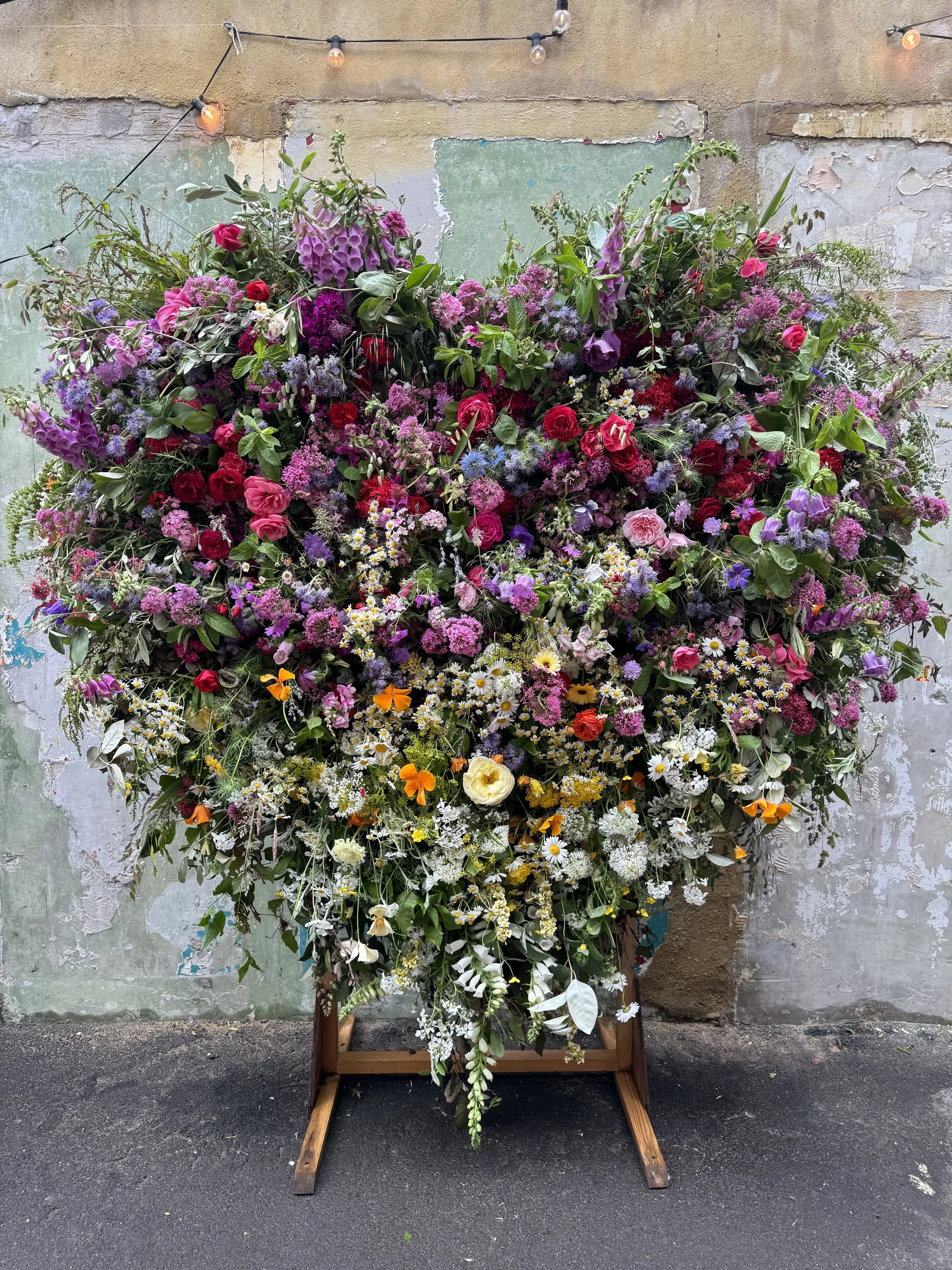 A large colorful floral arrangement with pink, purple, white, and yellow flowers displayed on a wooden stand against a worn wall.