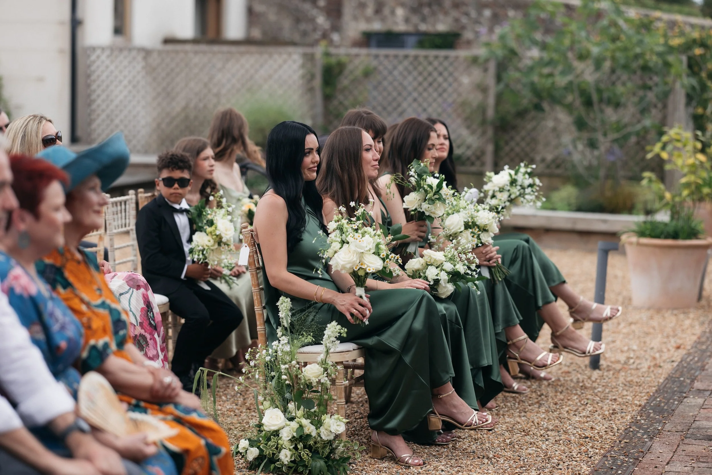 Group of women, dressed in green, sitting outdoors at a wedding, holding bouquets of white flowers. A young boy in a black tuxedo and sunglasses is also present among them. The setting is decorated with floral arrangements and surrounded by a garden with trees and plants.