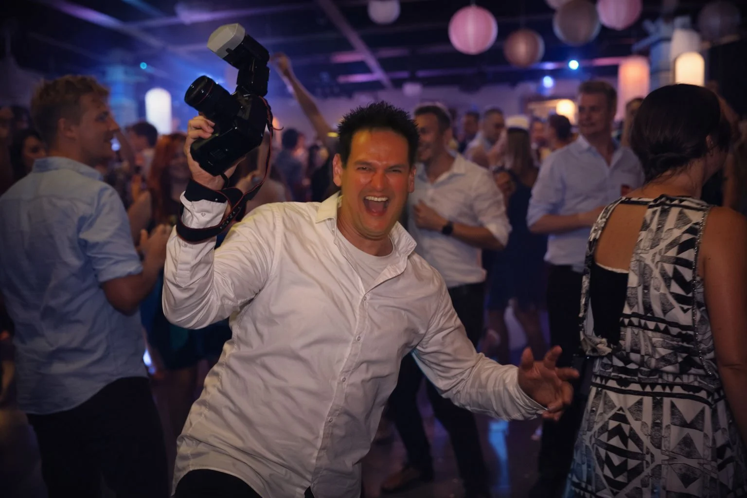 Man die danst en een camera vasthoudt in een feestzaal met andere mensen die ook dansen en plezier maken.