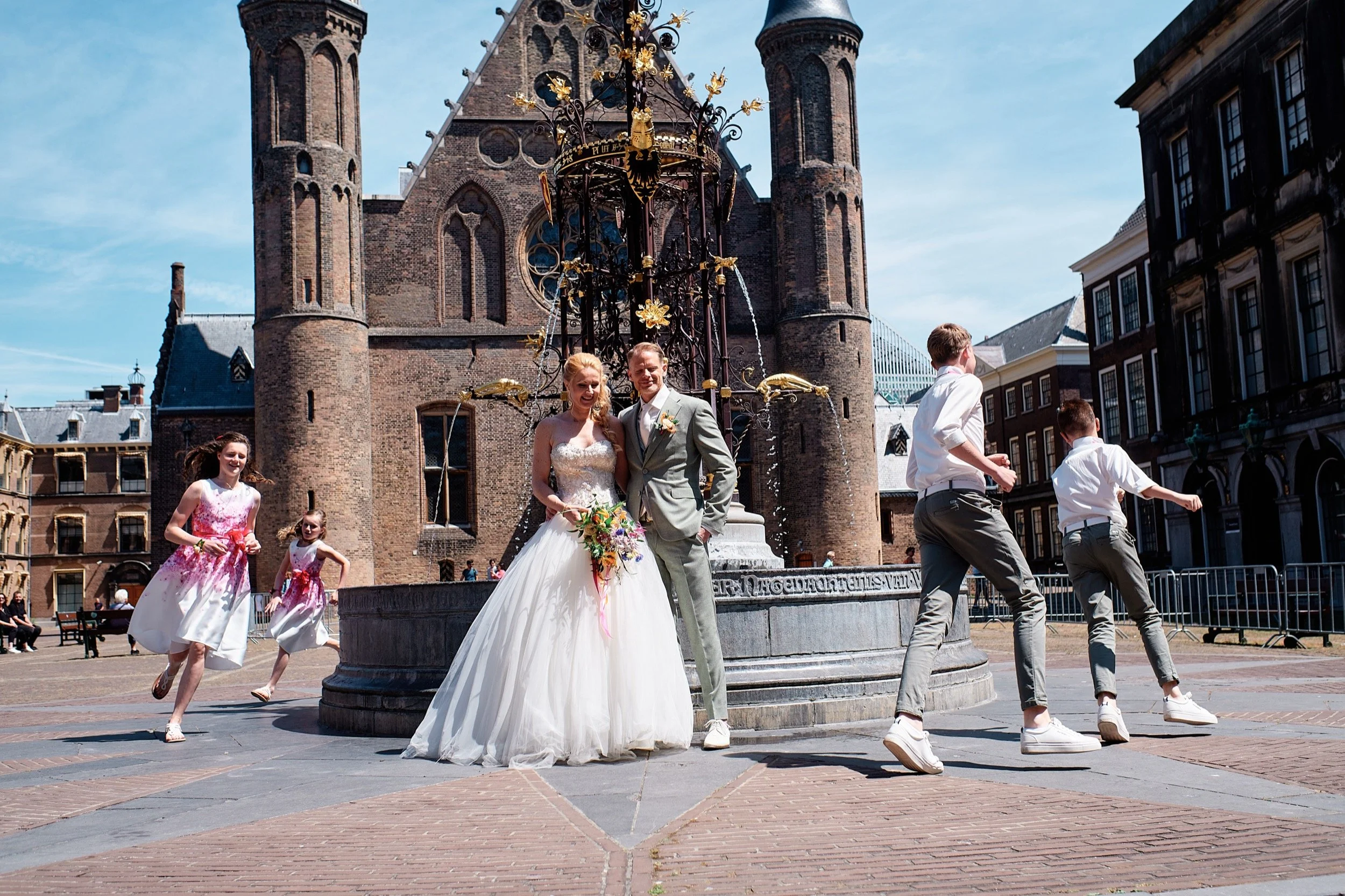 Een bruid en bruidegom poseert voor een fontein met kinderen die eromheen dansen op een zonnige dag in een historische stad.