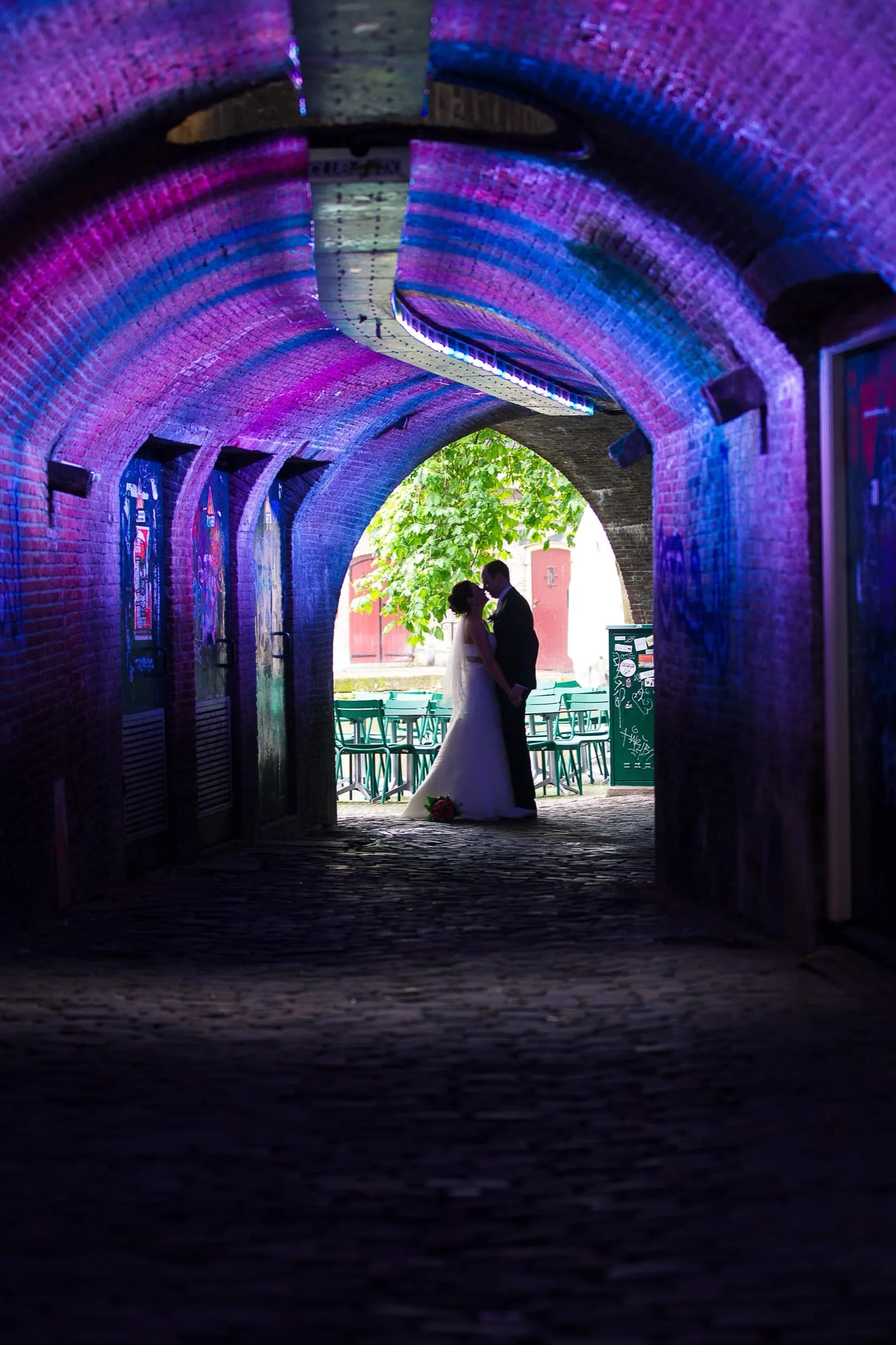 Bruid en bruidegom die elkaar kussen onder een kleurrijke overkapping, met buitenzicht op een groen park en stoelen in de achtergrond.