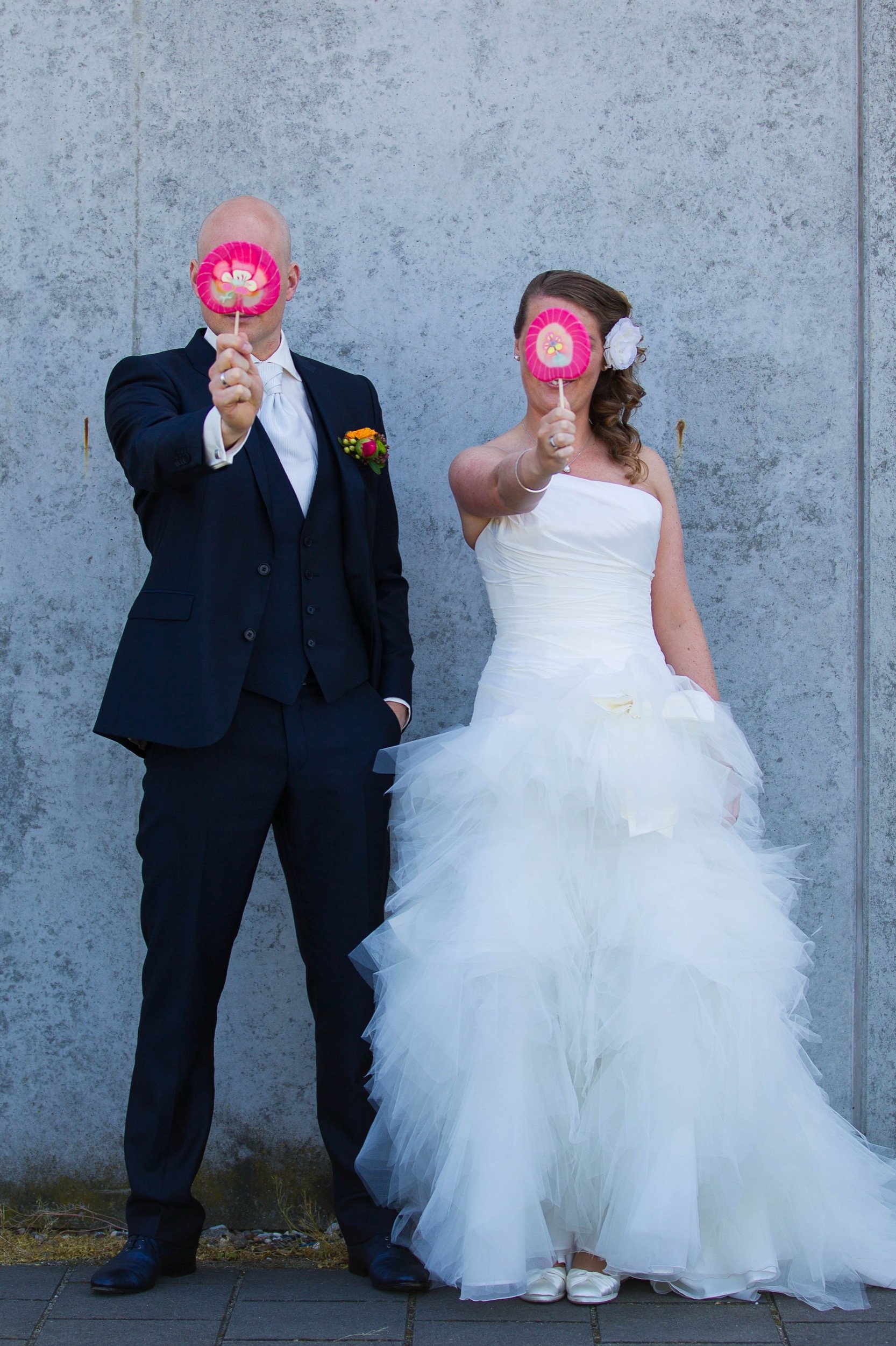 Een bruid en bruidegom staan naast elkaar voor een betonnen muur, beiden houden een roze lollipop voor hun gezicht die hun gezicht bedekt.
