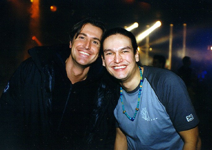 Twee mannen glimlachen en poseren voor de camera op een feest met donkere achtergrond en lichteffecten.