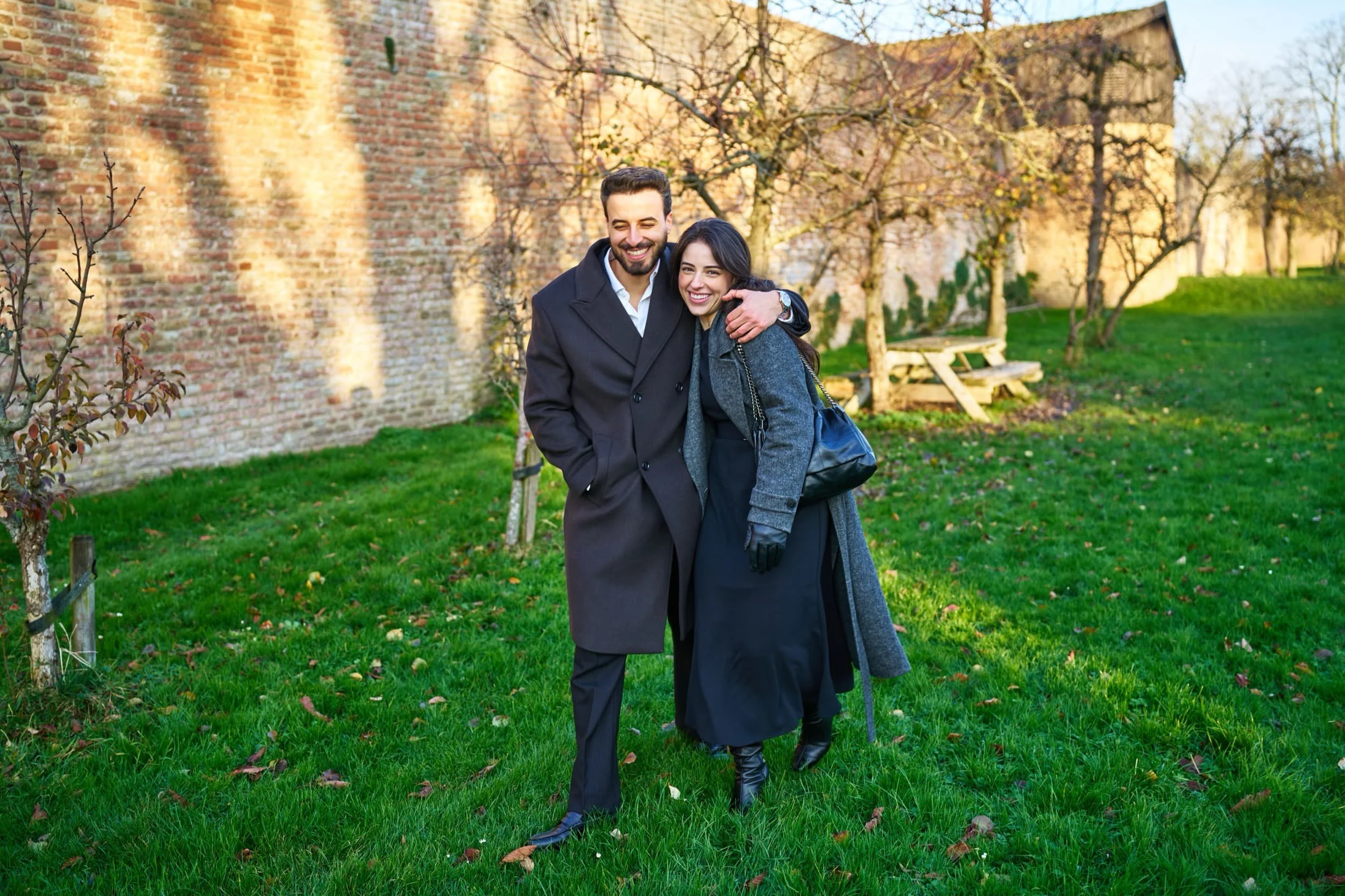 Een man en vrouw lachen en wandelen samen op een grasveld in de buitenlucht, omgeven door bomen en een oude bakstenen muur, tijdens een zonnige dag in de herfst.
