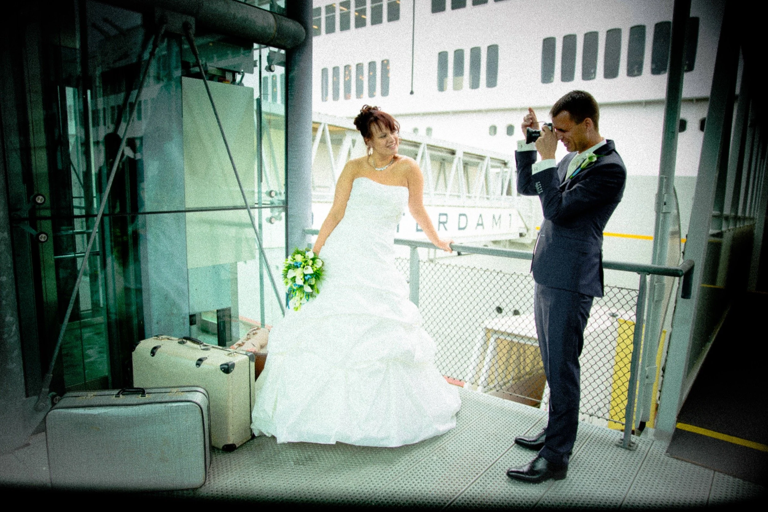 Een bruid in een witte trouwjurk poseren voor een fotograaf op een luchthaven, met koffers op de grond naast haar.