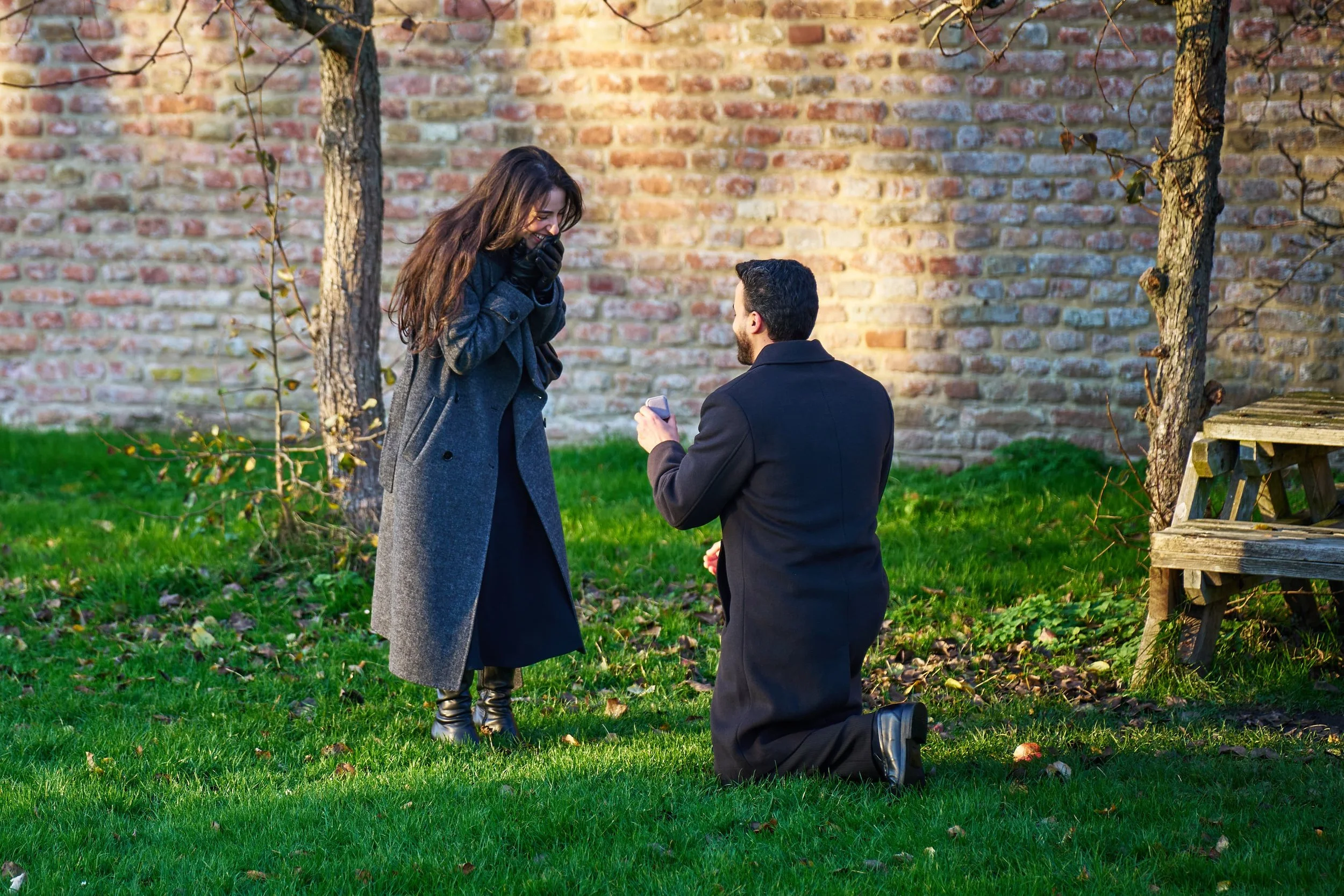 Man doet een voorstel aan een vrouw in een park, hij knielt op één knie met een ring in de hand, zij lijkt verrast en blij.