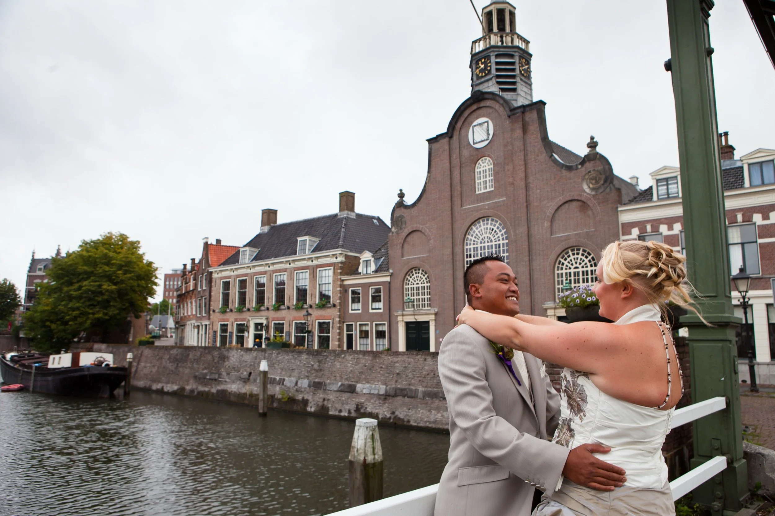 Een bruidspaar dat gelukkig samen danst op een brug over een kanaal in een historische stad, met oude gebouwen en een kerk op de achtergrond.