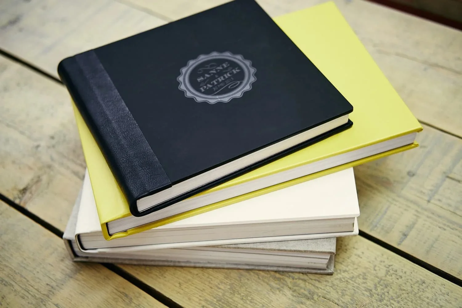 Een stapel boeken op een houten tafel, met de bovenste boek bedekt met een zwarte omslag met een logo en tekst, de tweede boek heeft een gele omslag, en de onderste boeken hebben witte en beige omslagen.