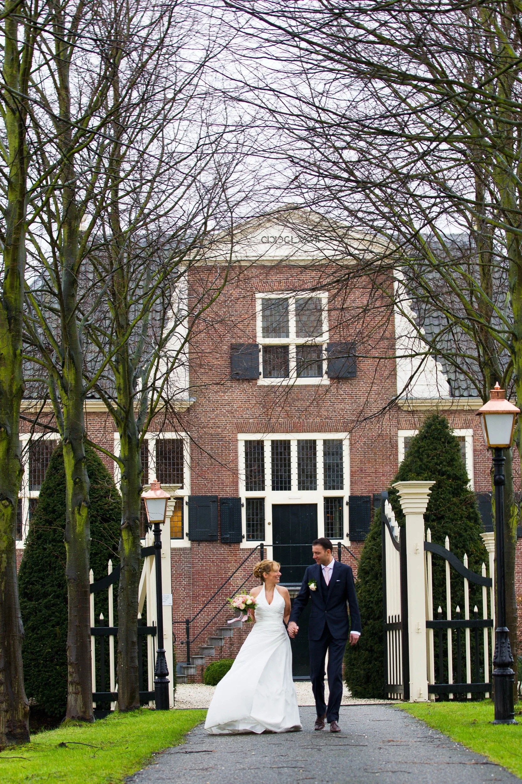 Een bruid in een witte jurk en een bruidegom in een donkere pak lopen samen hand in hand door een tuin naar een groot huis met een hek, bomen en lantaarns.