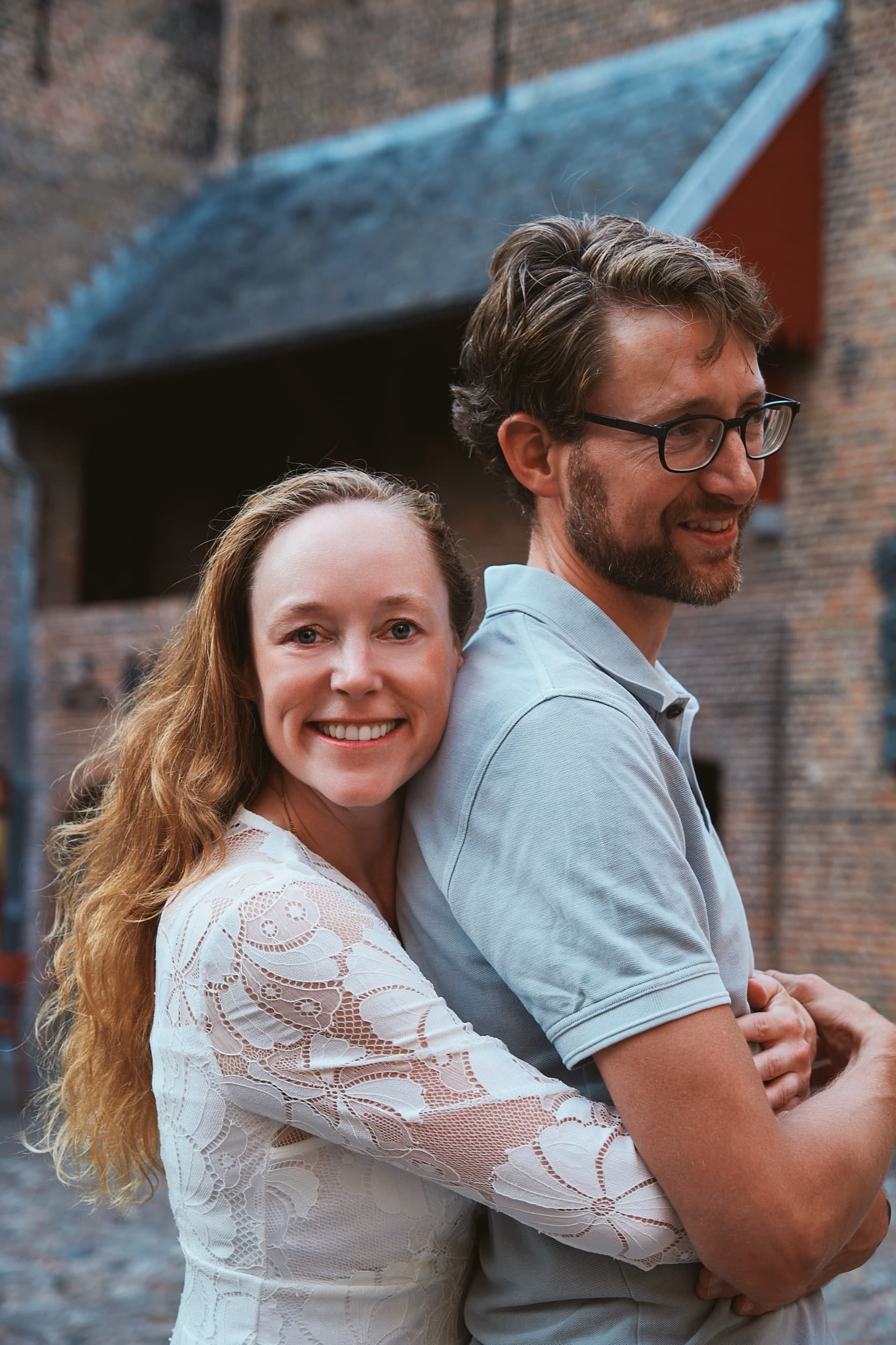 Een vrouw met lange, golvende, blonde haren en een witte kanten blouse, omhelst een man met een baard, bril en lichtblauwe polo, buiten bij een oude bakstenen structuur.