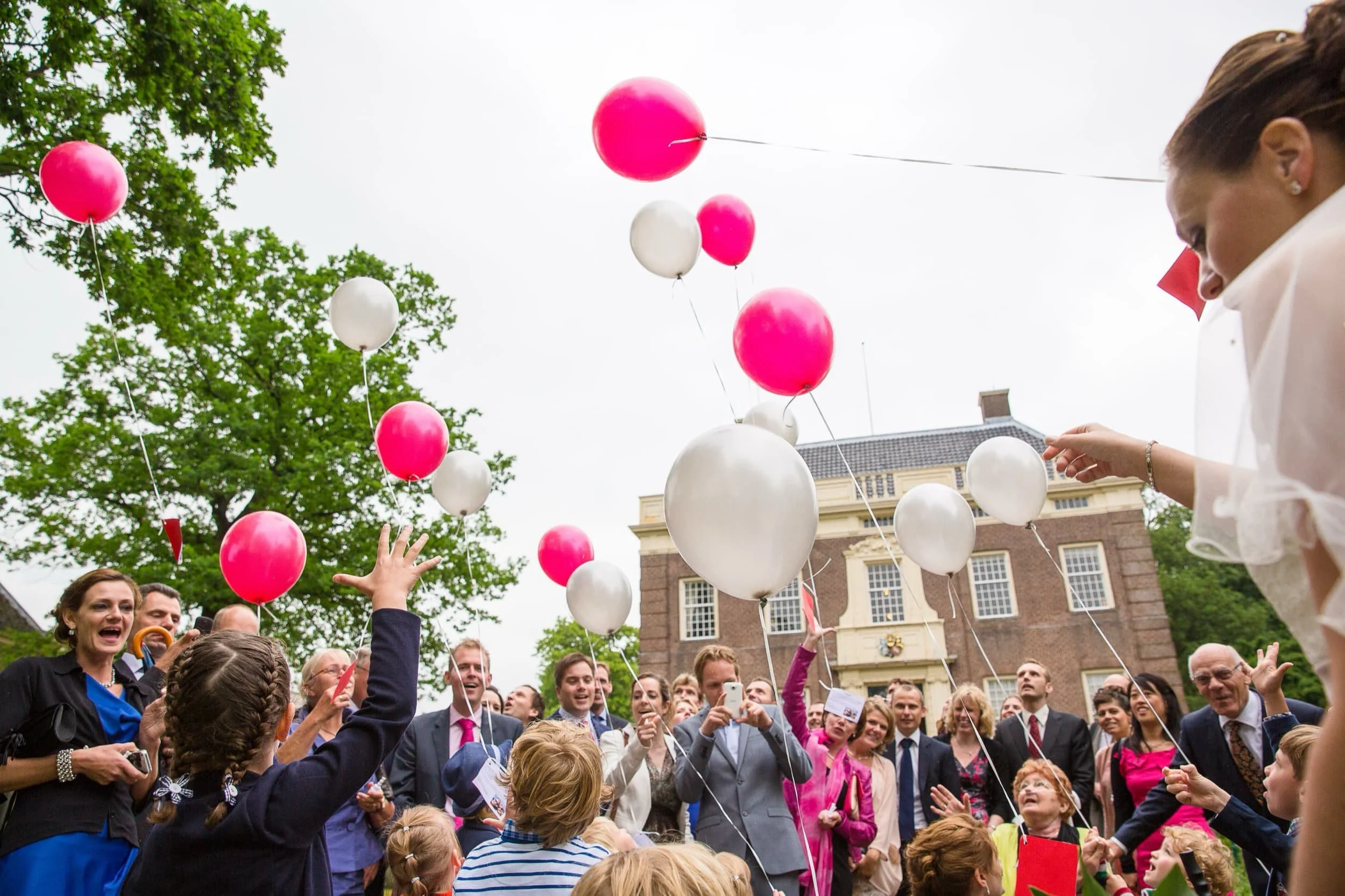Mensen vieren buiten tijdens een feest, met roze en witte ballonnen in de lucht en blije gezichten.