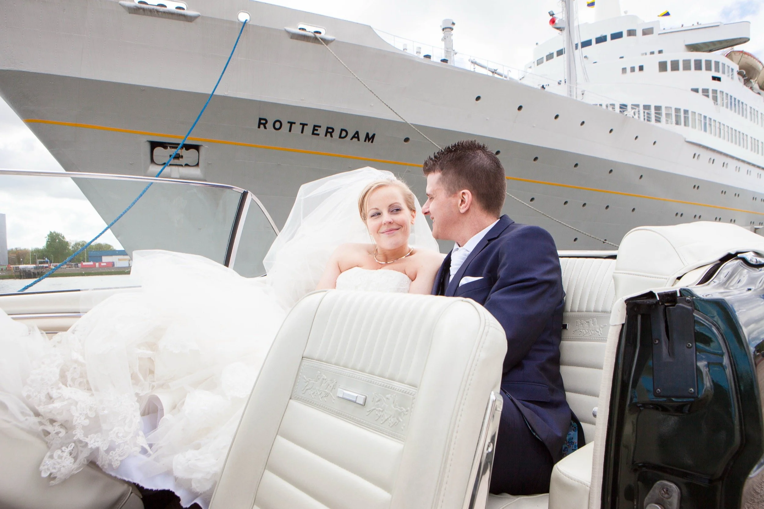 Een bruid en bruidegom op een luxe boot met een grote cruiseschip op de achtergrond. Ze zitten dicht bij elkaar en kijken elkaar liefdevol aan.