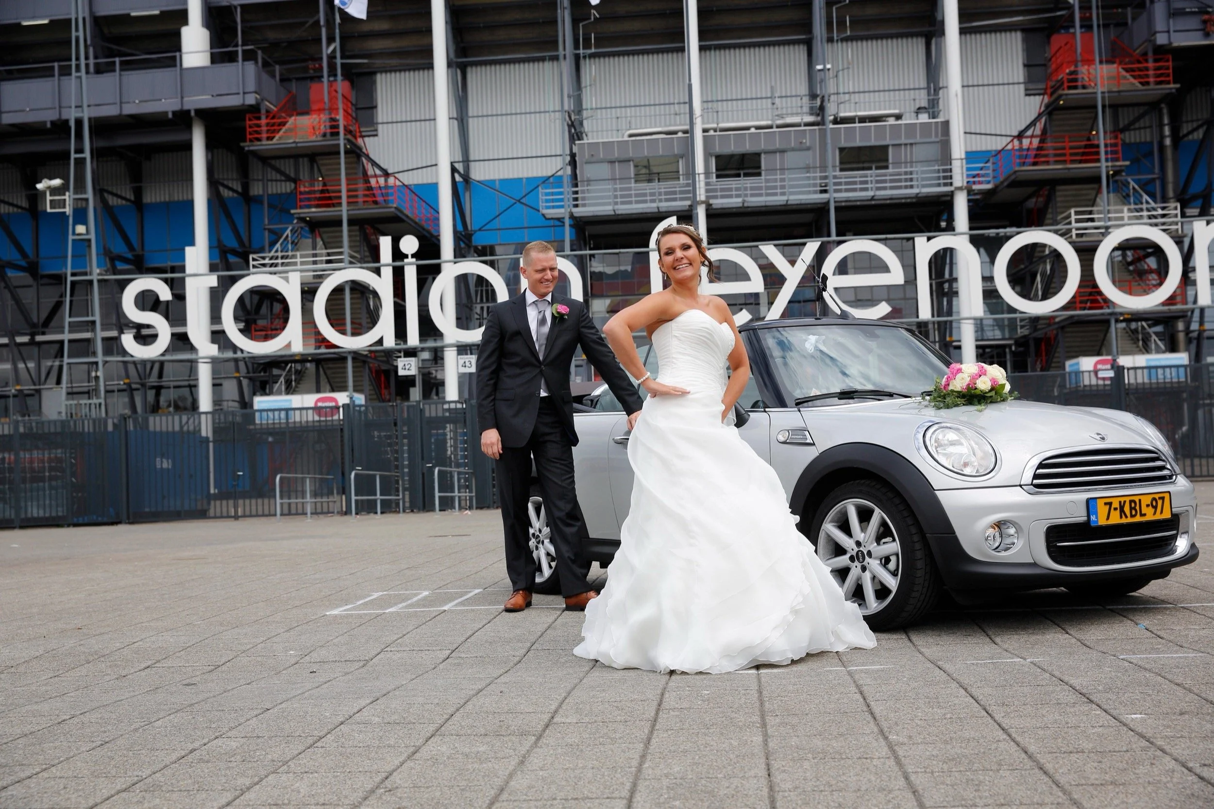 Een bruid en bruidegom poseert naast een kleine witte auto met bloemen op de motorkap voor een stadion met een groot 'stadion' bord op de achtergrond.