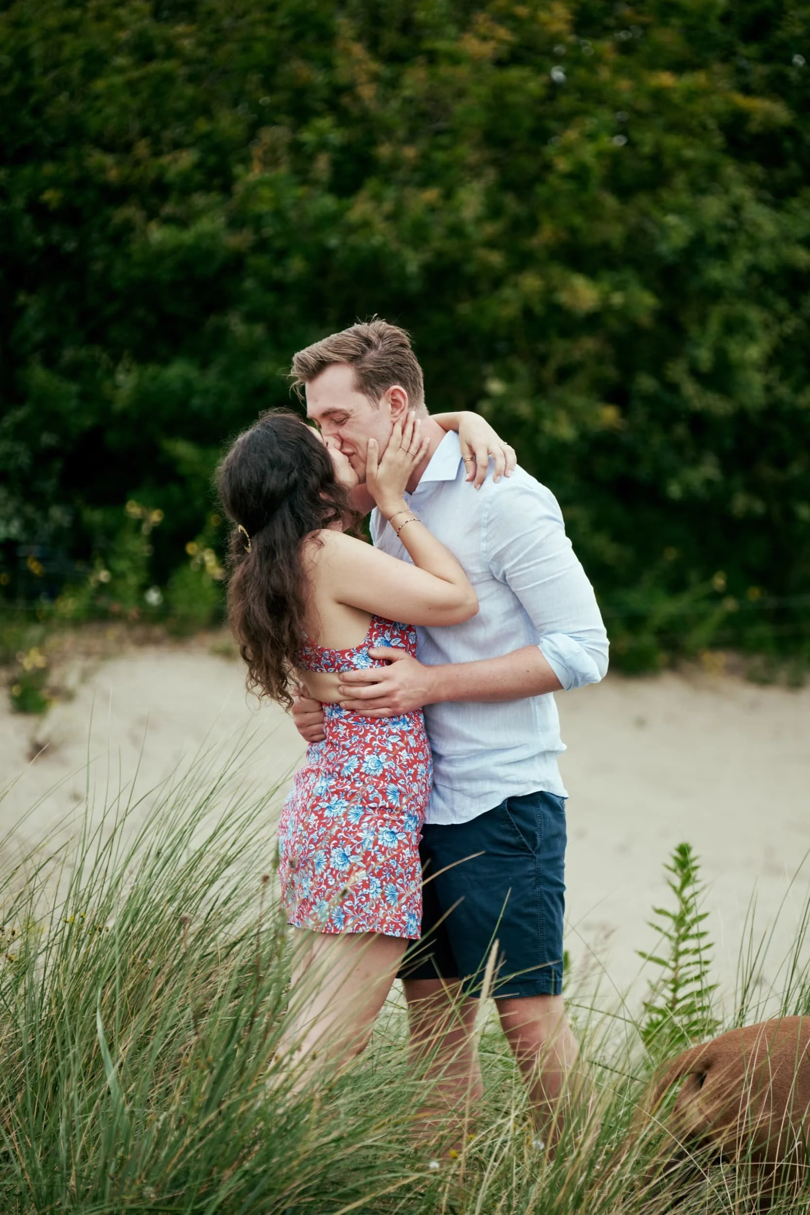 Een liefdevol koppel kust elkaar terwijl ze op het strand staan, omringd door gras en struiken, met een groene bos op de achtergrond.