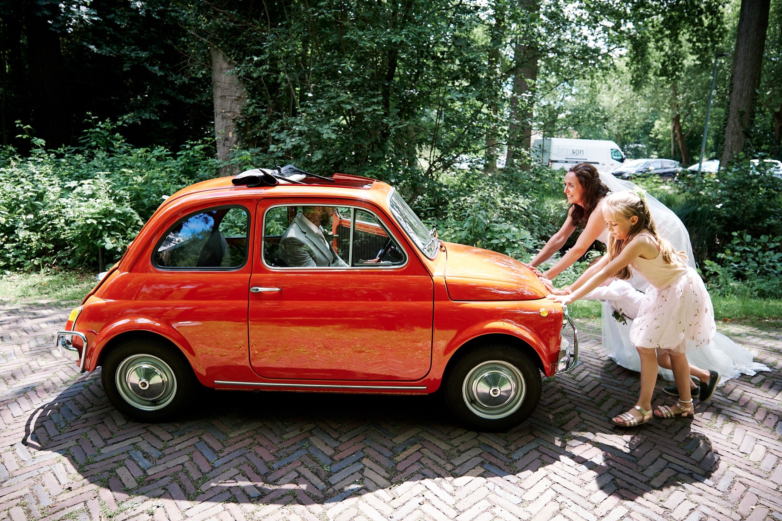 Twee vrouwen en een meisje duwen een kleine, rode, vintage auto op een bosweg.