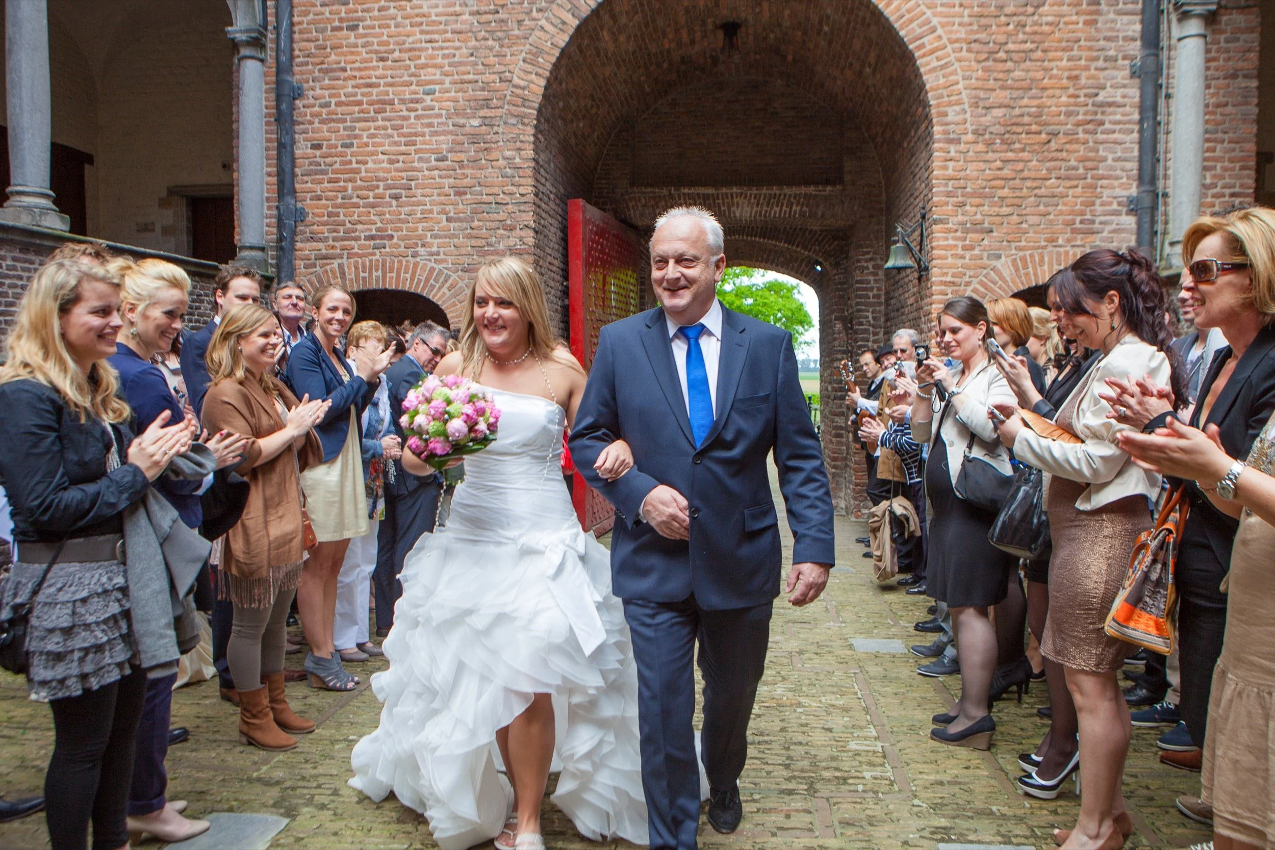 Bruid en vader lopen door een menigte van gasten die applaudisseren en foto's maken, buiten een historische bakstenen poort.