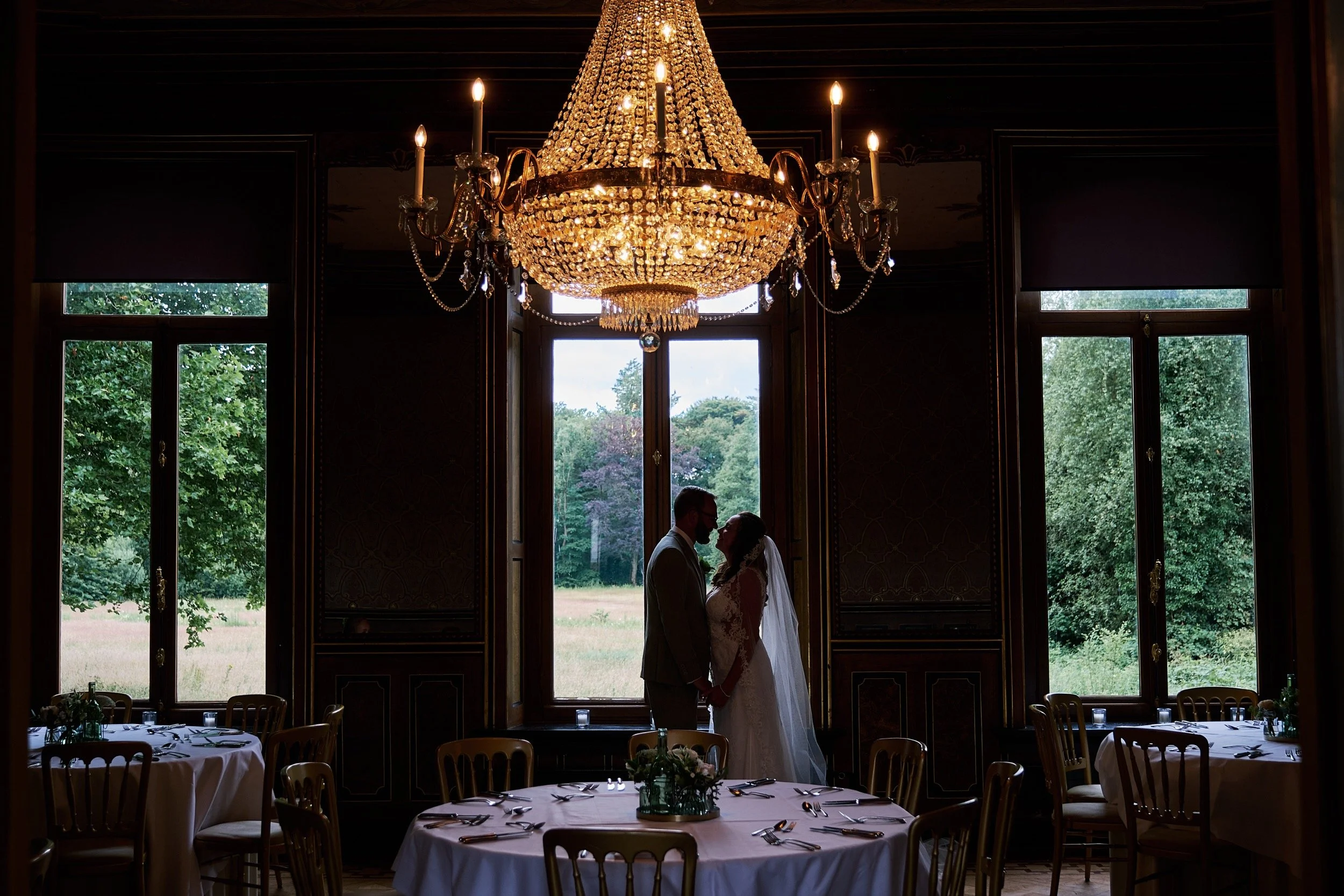 De silhouetten van een bruid en bruidegom die elkaar in een kamer met grote ramen en een chandelier vasthouden, met uitzicht op een groene tuin.