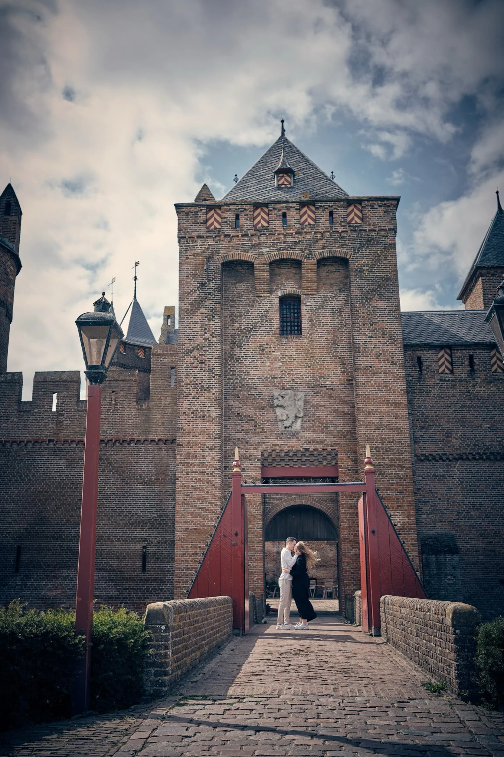 Twee mensen stonden onder de poort van een middeleeuws kasteel, omarmd met de recente wind in hun haren, terwijl de kasteel torent boven hen uit tegen een bewolkte hemel.