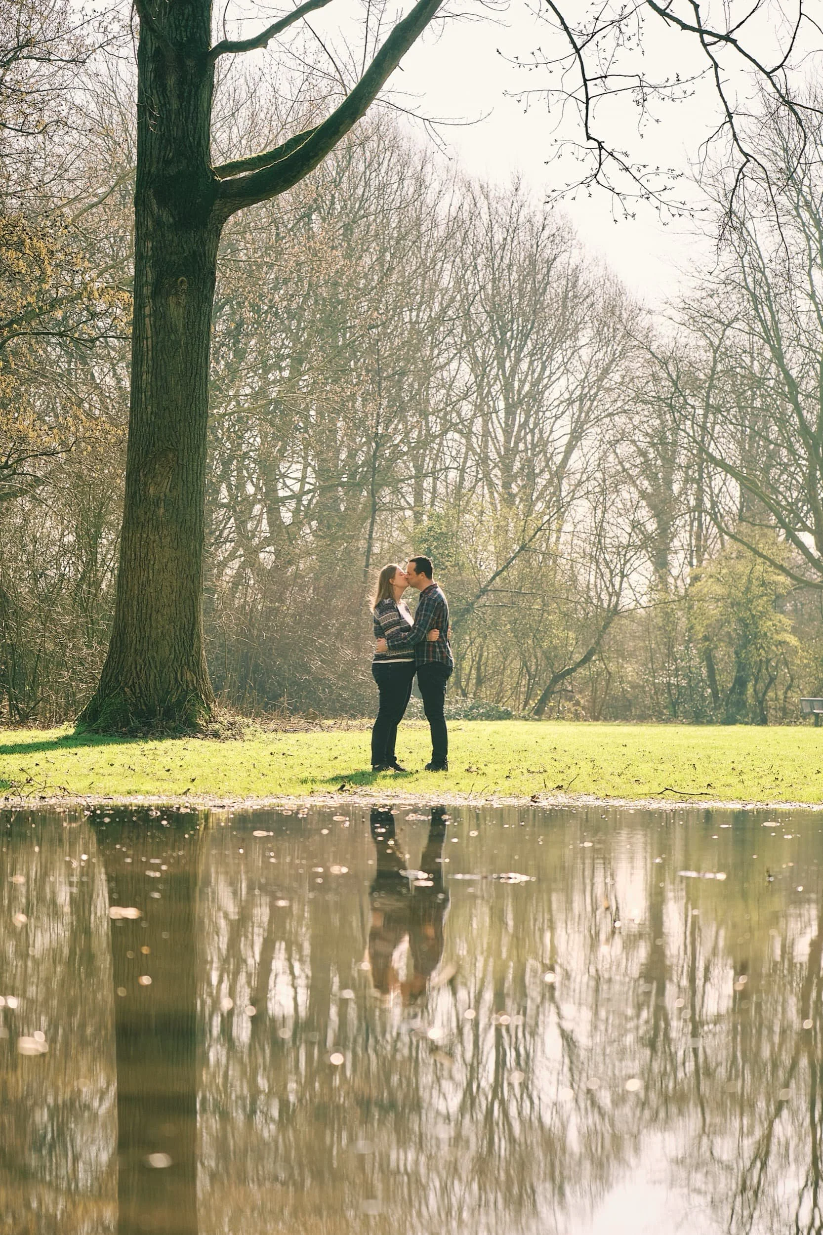 Een man en vrouw kussen elkaar in een park met een grote boom, herfstbladeren en een waterpartij die hun reflectie toont.